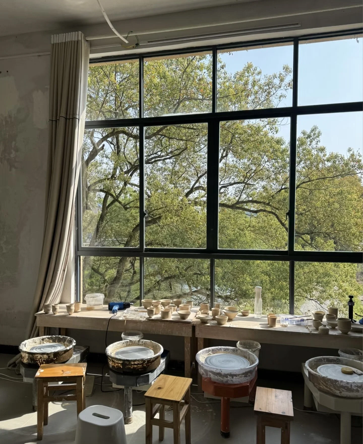 A pottery studio with a large window showing green trees outside. The windowsill has numerous pottery pieces in progress, along with pottery wheels, stools, and tools.
