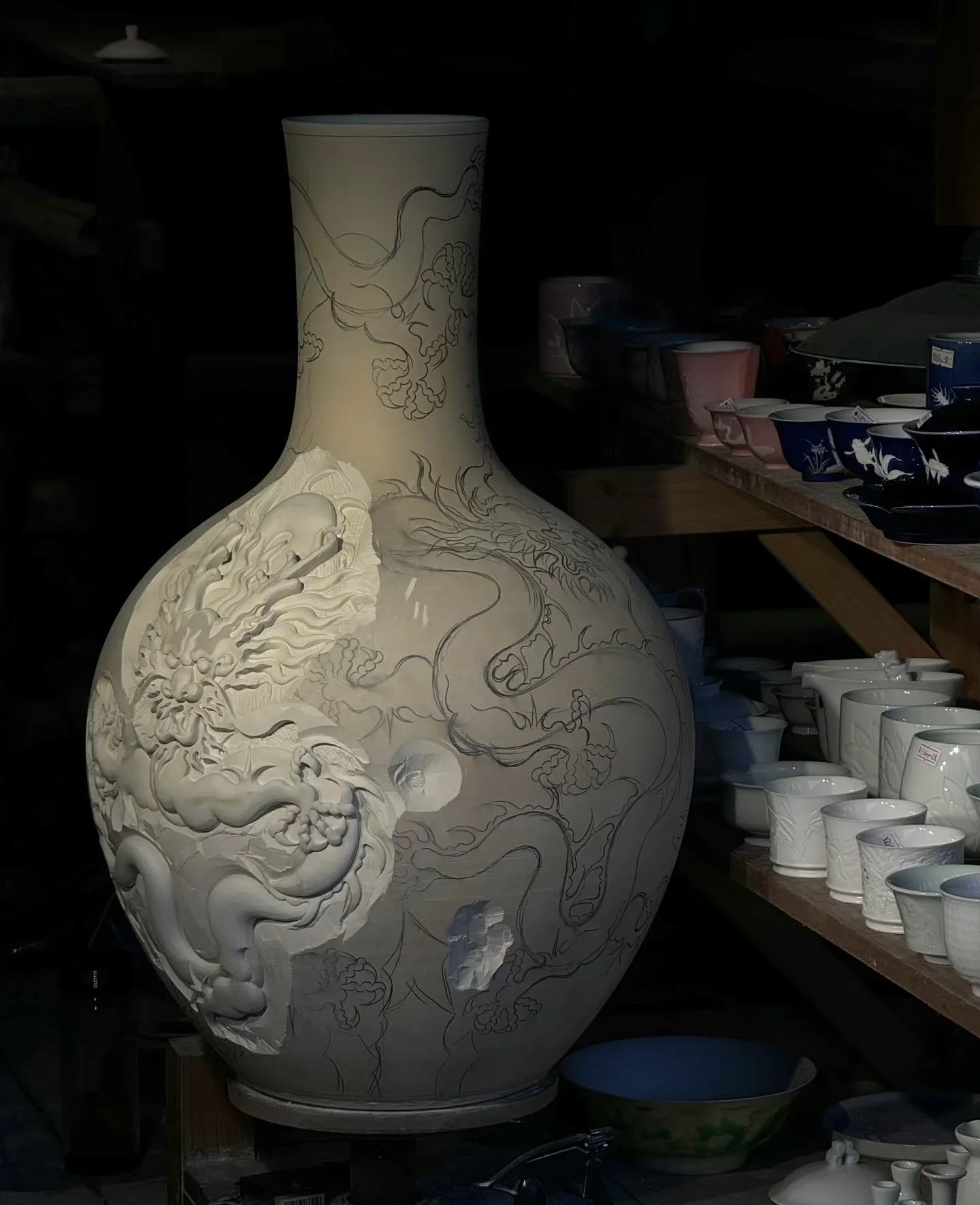 Large ceramic vase with carved dragon and cloud designs in an antique shop, surrounded by various cups and bowls.