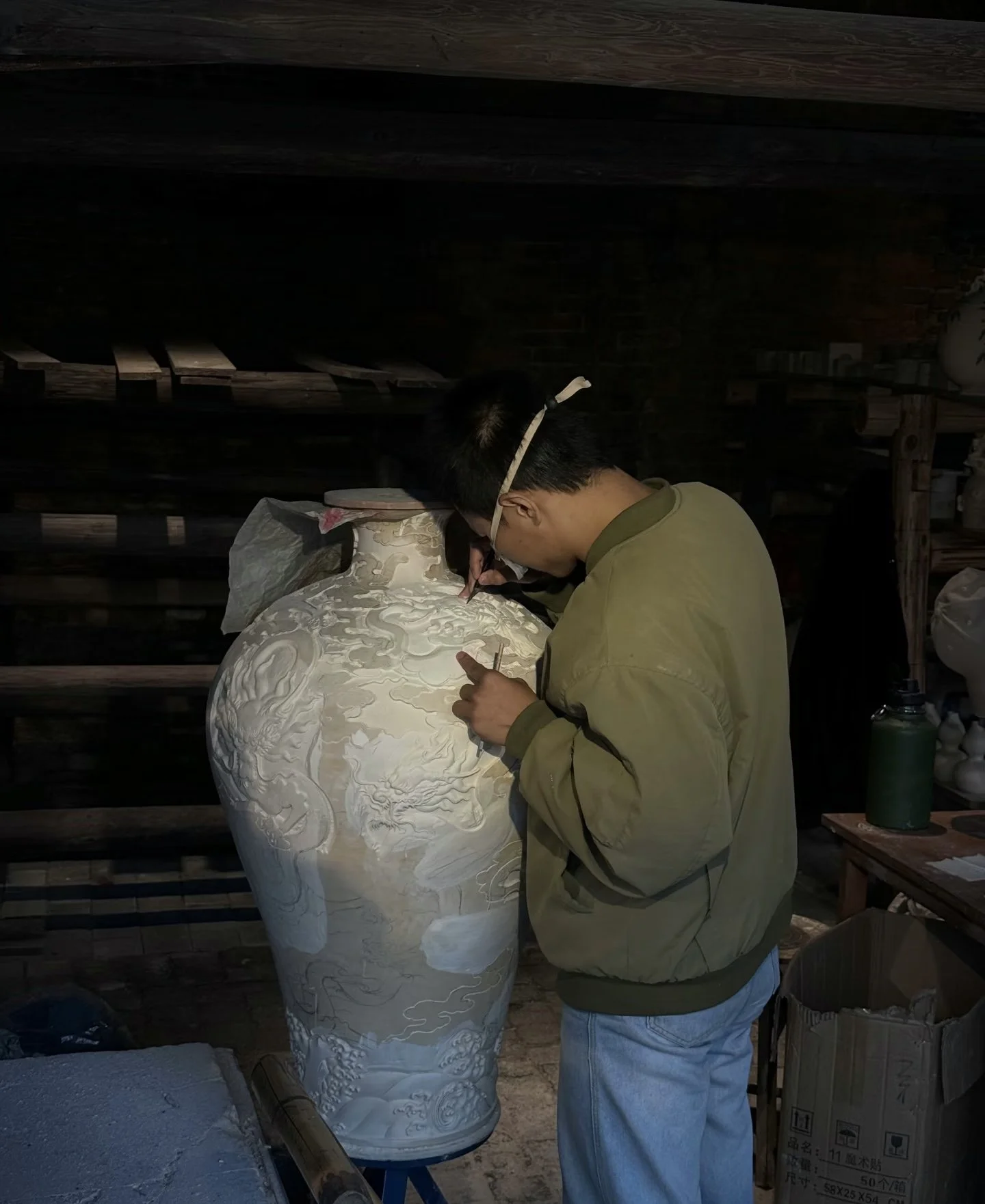 A person wearing glasses works on a large, intricately carved white ceramic vase, using a fine tool, in a workshop with wooden shelves and pottery items.