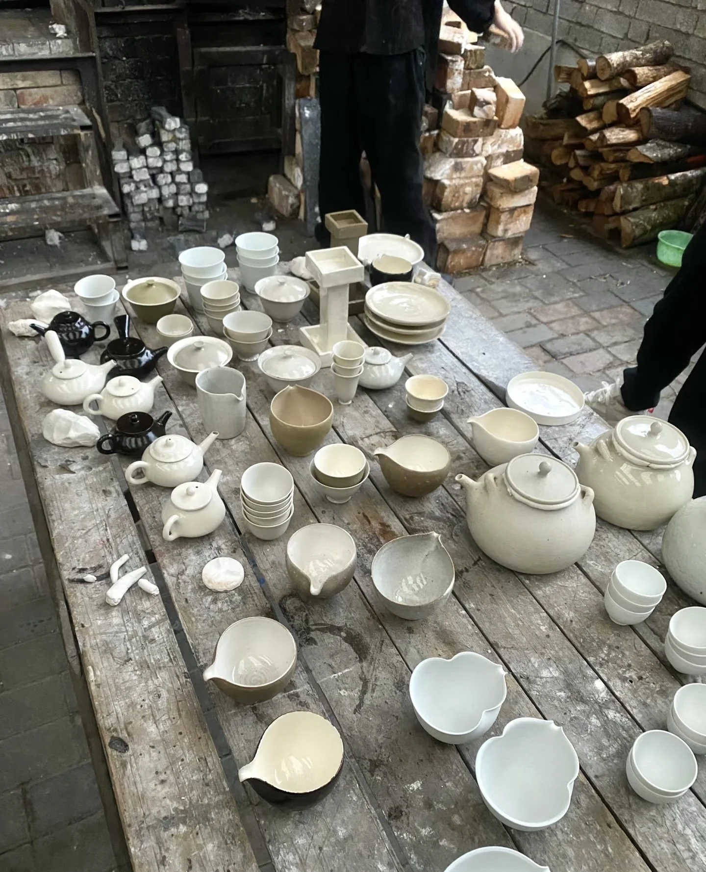 A rustic wooden table covered with various unfinished ceramic pottery pieces, including teapots, bowls, cups, and vases in neutral colors. In the background, there are stacked logs and bricks, with two people partially visible standing nearby.
