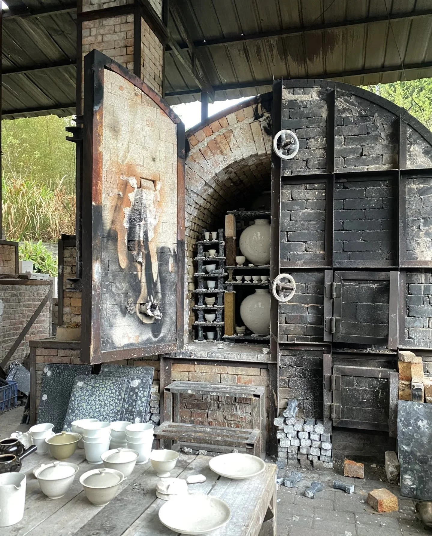 An outdoor kiln or furnace with a fireproof chamber, blackened inside, containing shelves of white ceramic pots and bowls, some on a small shelf, with unfinished ceramic pieces on the ground and a table of completed ceramics in front.