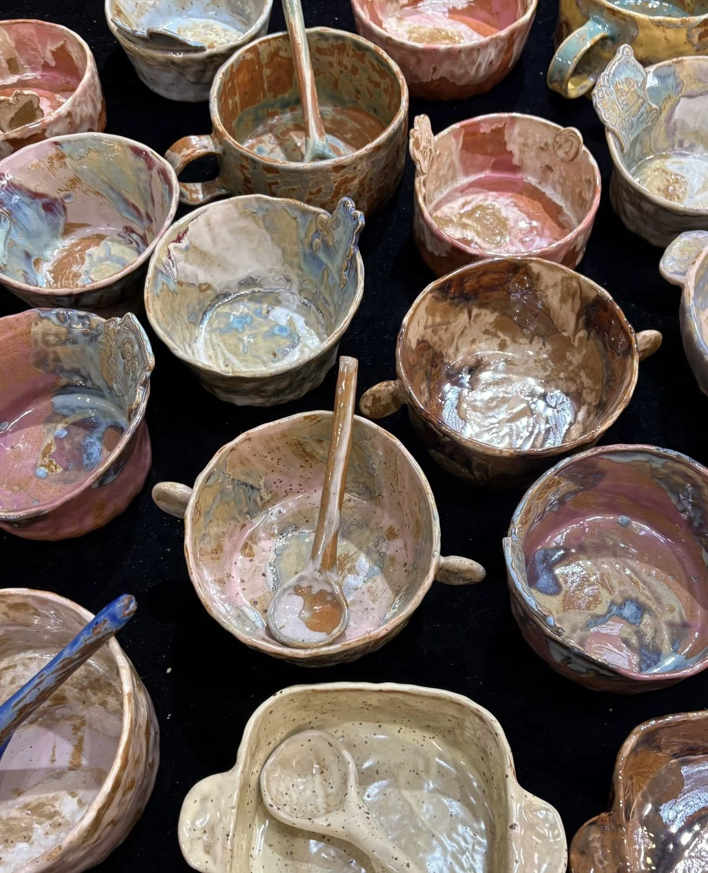 A collection of colorful, handcrafted ceramic bowls and cups with spoons inside some of them, displayed on a black surface.