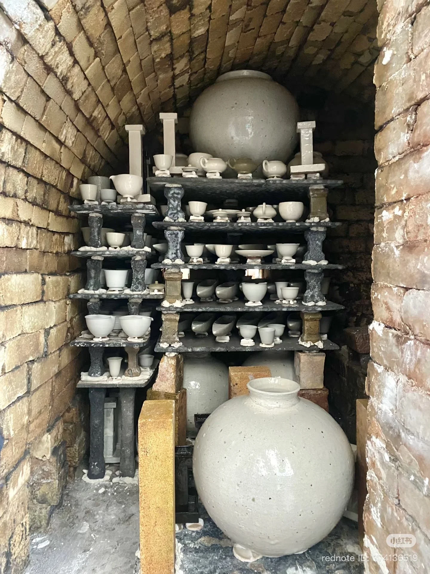 Storage alcove with brick walls and a curved brick ceiling, containing several shelves with multiple white ceramic bowls, cups, and small dishes, along with two large round ceramic jars at the top and bottom.