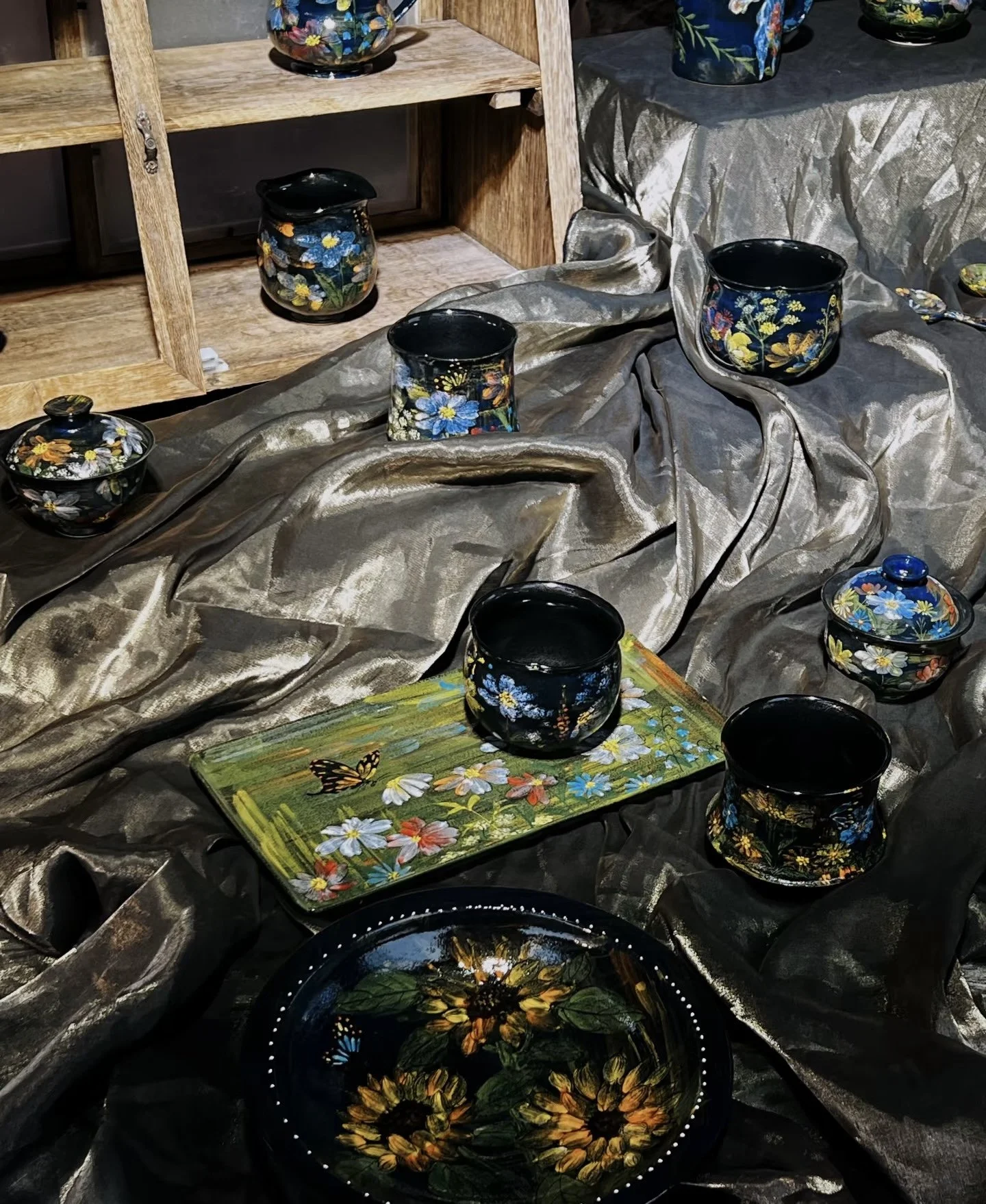 Display of hand-painted floral ceramics, including bowls and cups, arranged on satin fabric with a wooden shelf in the background.