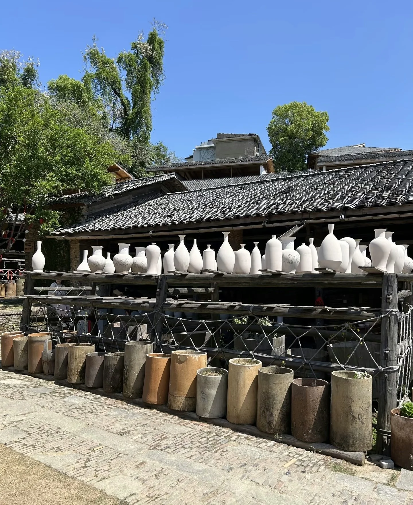 Outdoor display of various unglazed ceramic vases and pots on wooden shelves and beneath, in front of traditional buildings with tiled roofs and trees under a clear blue sky.