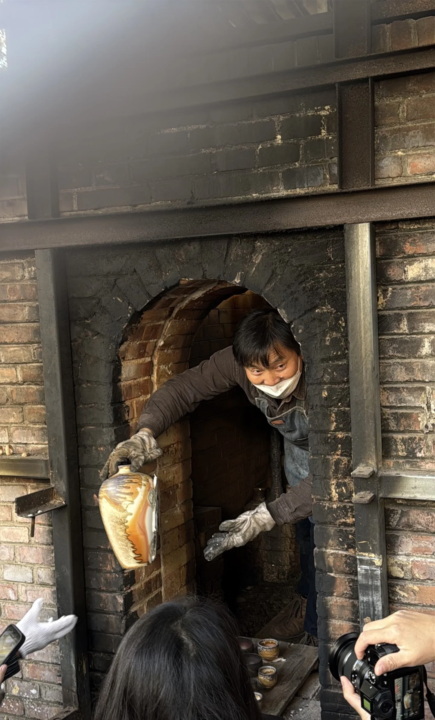 Man with gloves and face mask placing decorative objects into a brick oven surrounded by a group of people, some taking photos.