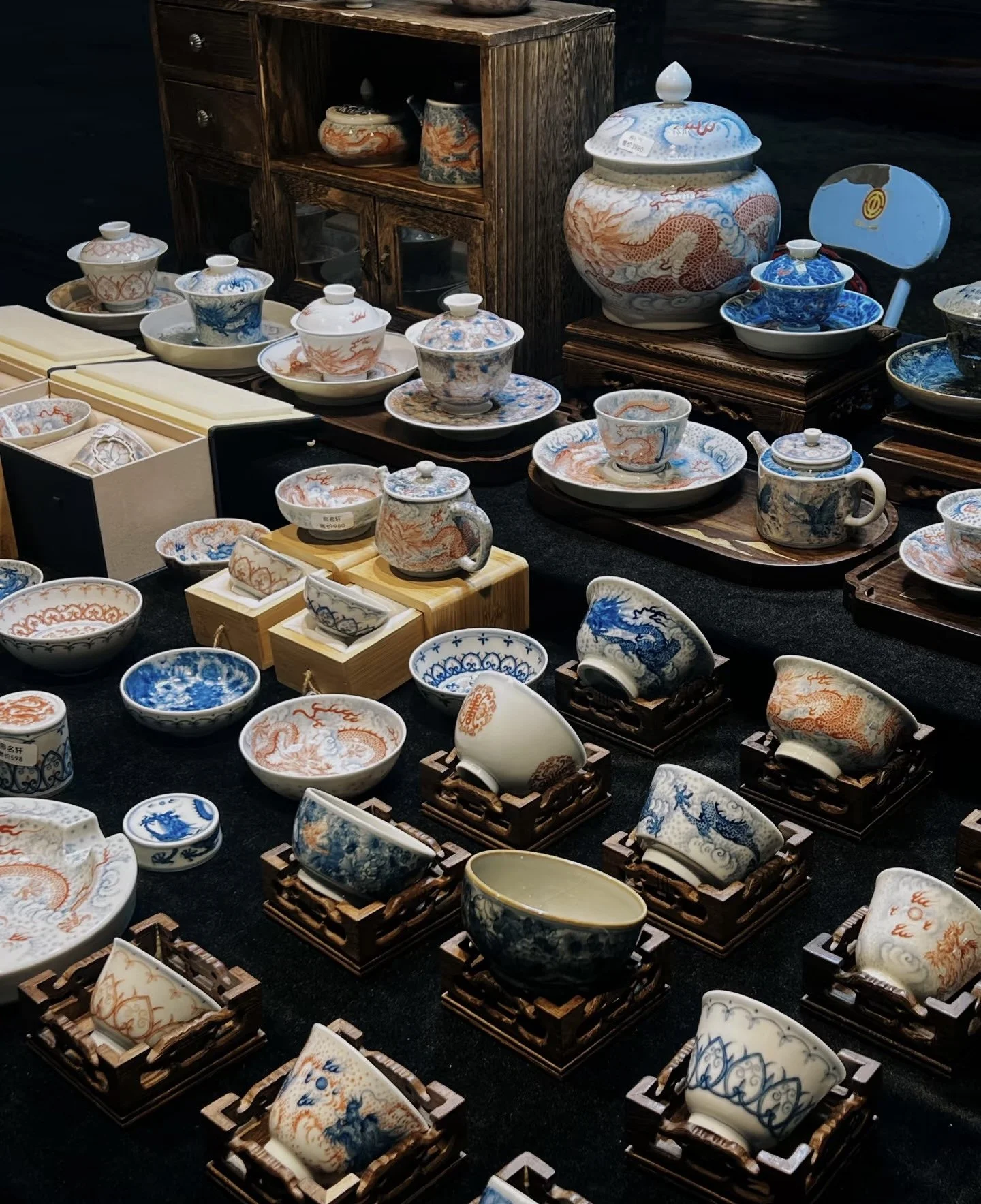 A display of various Chinese-style porcelain bowls, teapots, and dishes with blue and red dragon motifs, arranged on black fabric with some wooden stands and boxes.