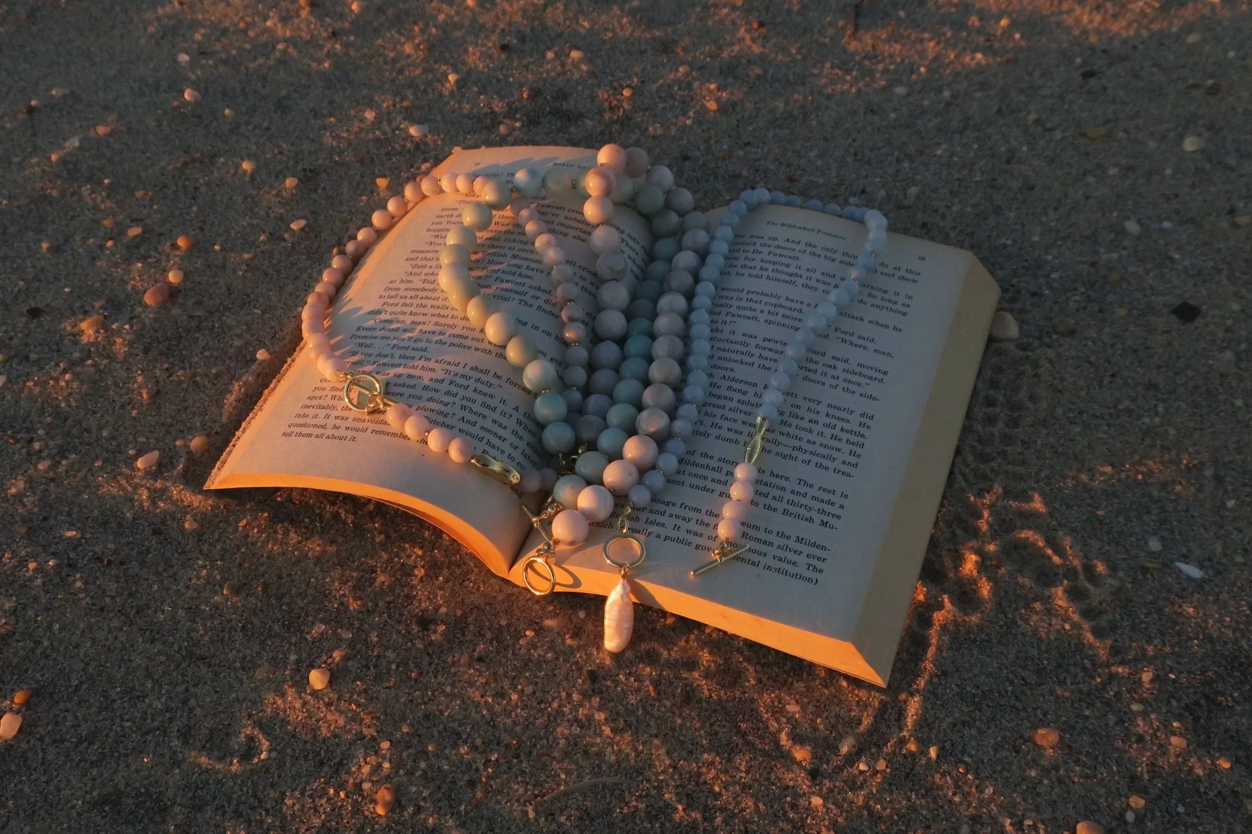 An open book at the beach with Lomi necklaces between the pages.