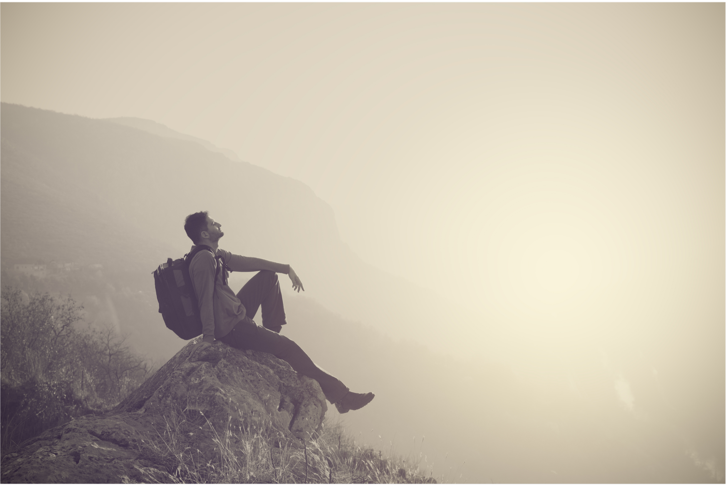A man with a backpack sitting on a rock, overlooking the landscape, during sunset or sunrise.