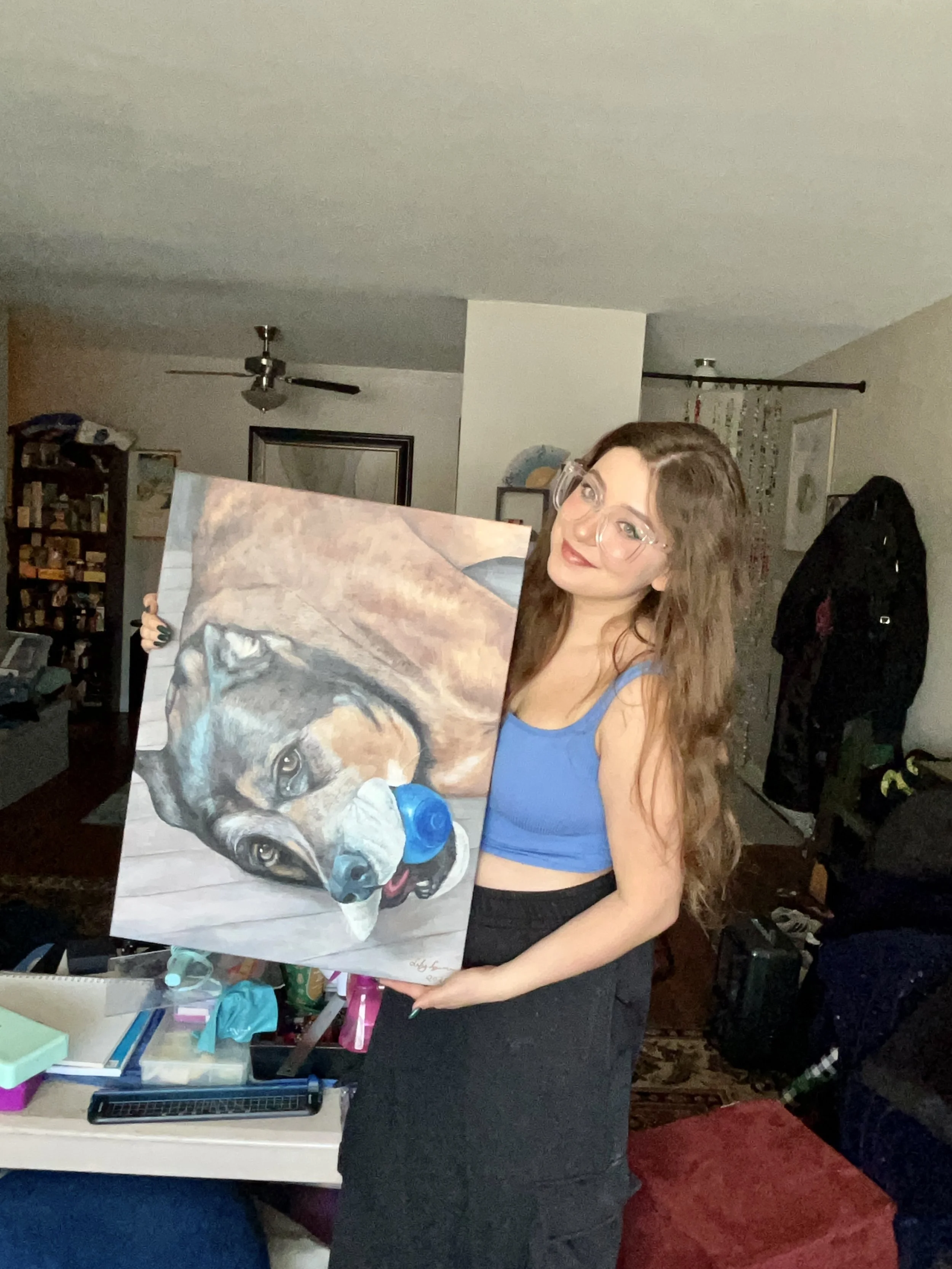 A woman with long, wavy brown hair and glasses wearing a blue sleeveless top and black skirt holding a painting of a dog with a blue ball in its mouth inside a living room.