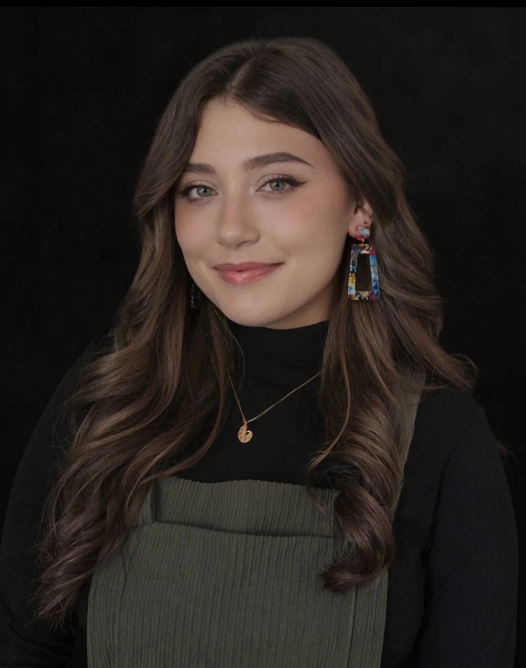 A young woman with long, wavy brown hair and blue eyes, smiling softly, wearing a black turtleneck, green overalls, colorful earrings, and a gold necklace against a black background.