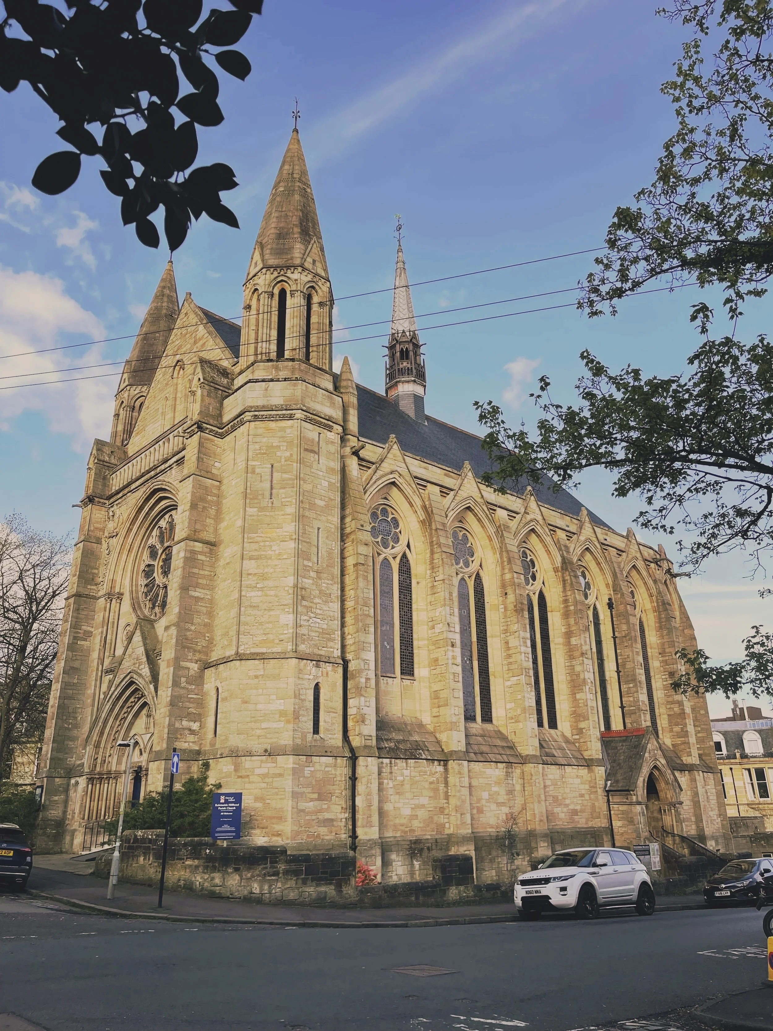 The stone Gothic-style exterior of Kelvinside Hillhead, with tall spires and stained glass windows, situated on a street with cars and trees.