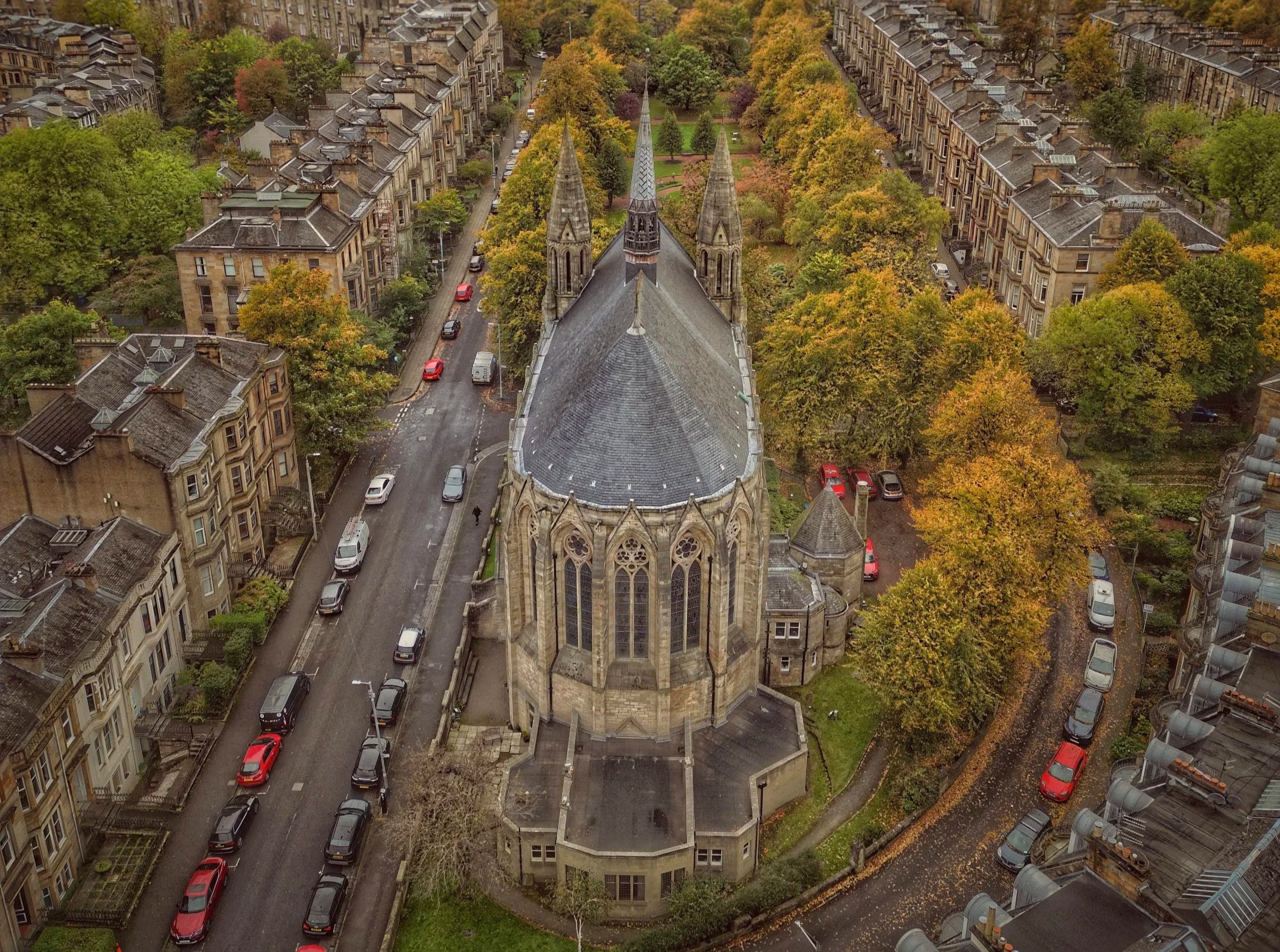 An aerial view of a historic stone church with a pointed steeple, surrounded by residential buildings and tree-lined streets with colorful autumn foliage.