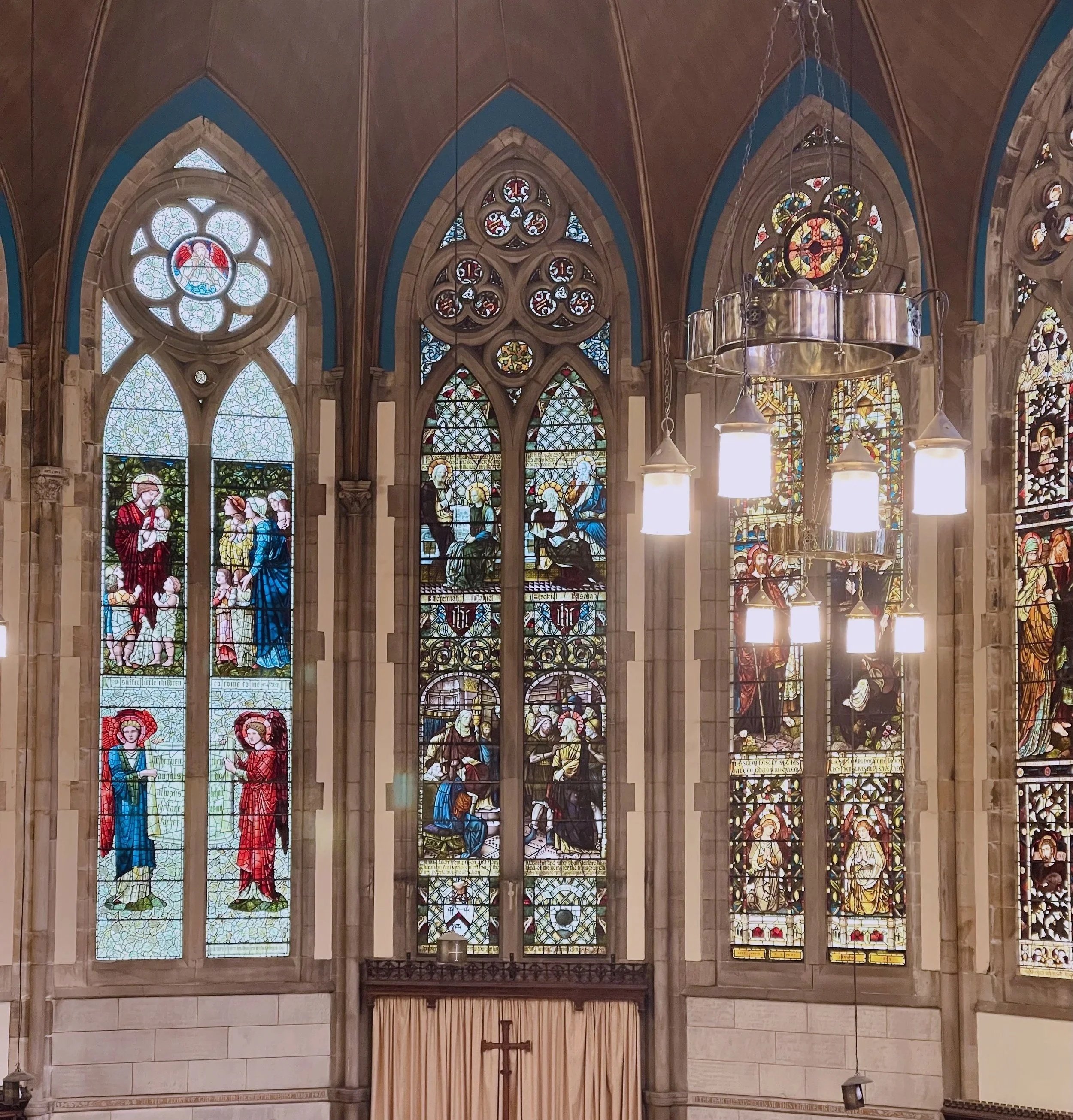 Interior of Kelvinside Hillhead church with stained glass windows depicting religious scenes and figures, a chandelier with lights, and a cross on the altar.