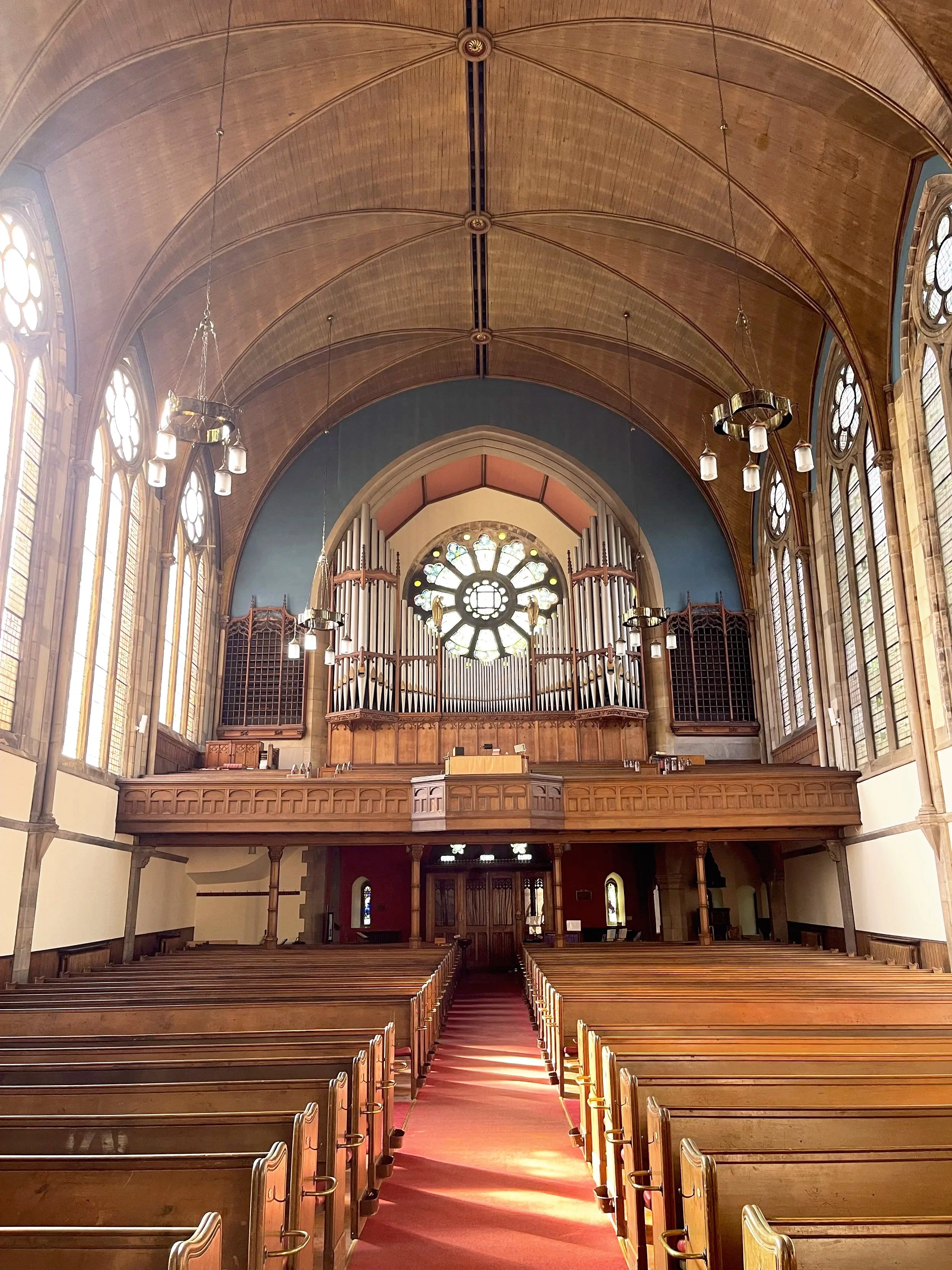 Interior of Kelvinside Hillhead church with wooden pews, stained glass windows, a pipe organ, and a large stained glass rose window at the front.