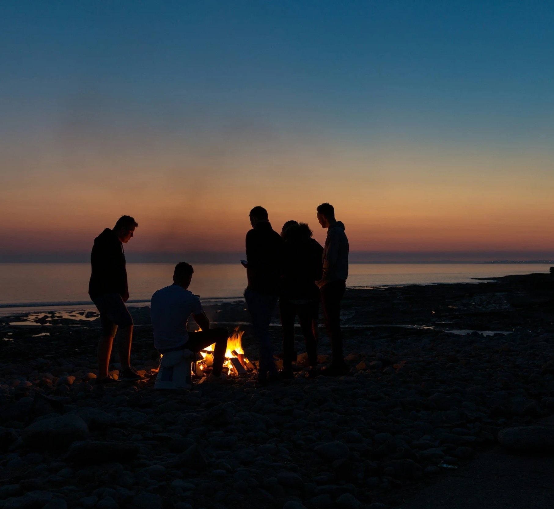 Silhouettes of people gathered around a small campfire on a rocky beach at sunset, with a calm ocean in the background and colorful sky overhead.