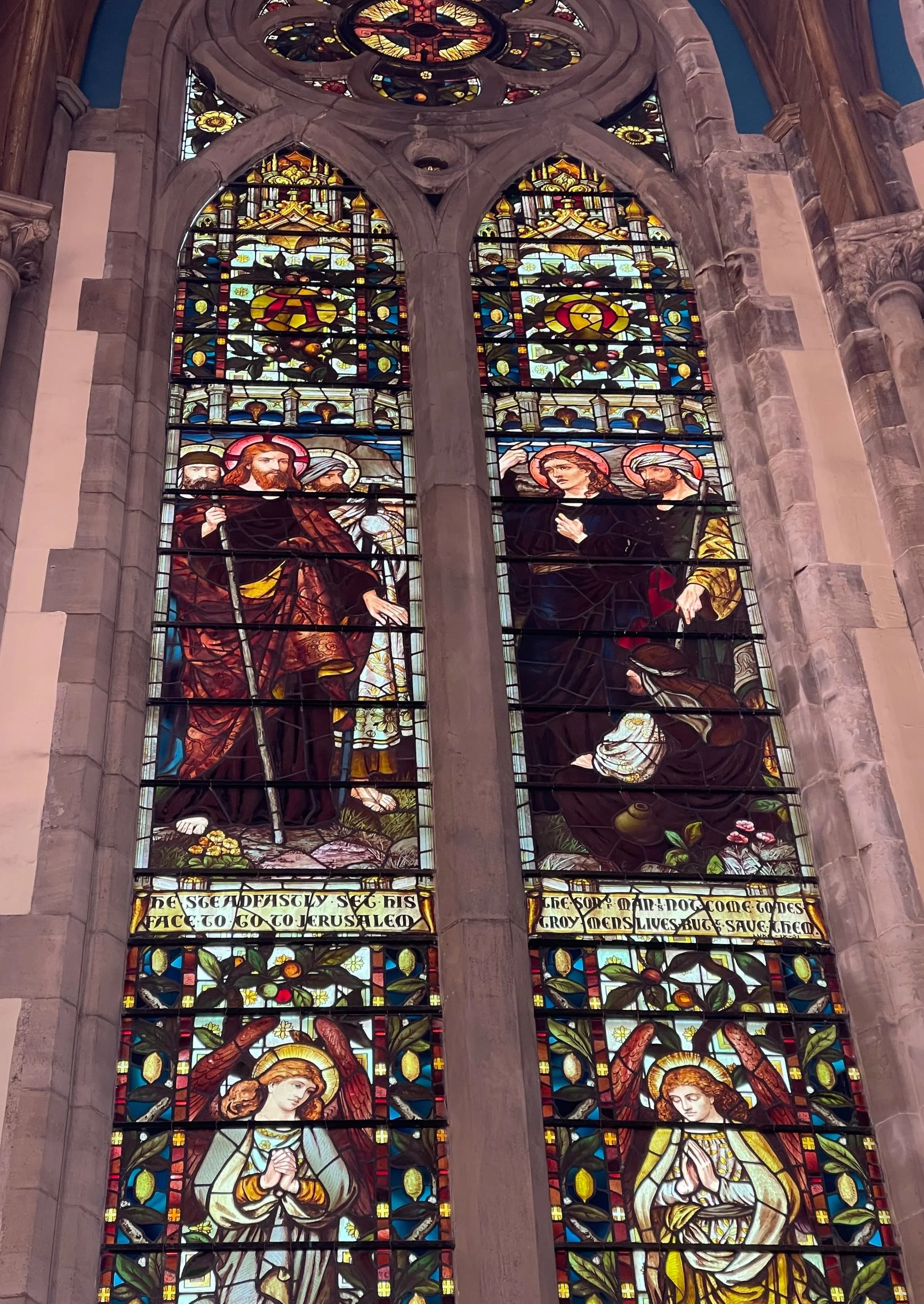 Colorful stained glass church window depicting Jesus Christ with two women, angels at the bottom, and various decorative patterns.