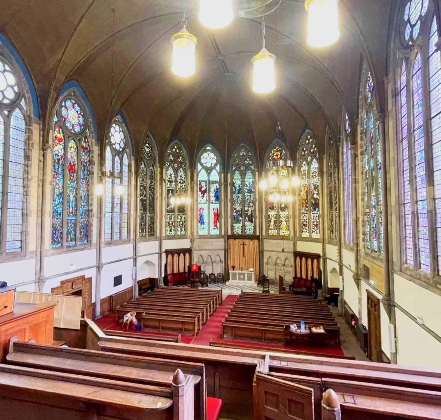 The interior of Kelvinside Hillhead, with stained glass windows, wooden pews, and an altar with a cross.
