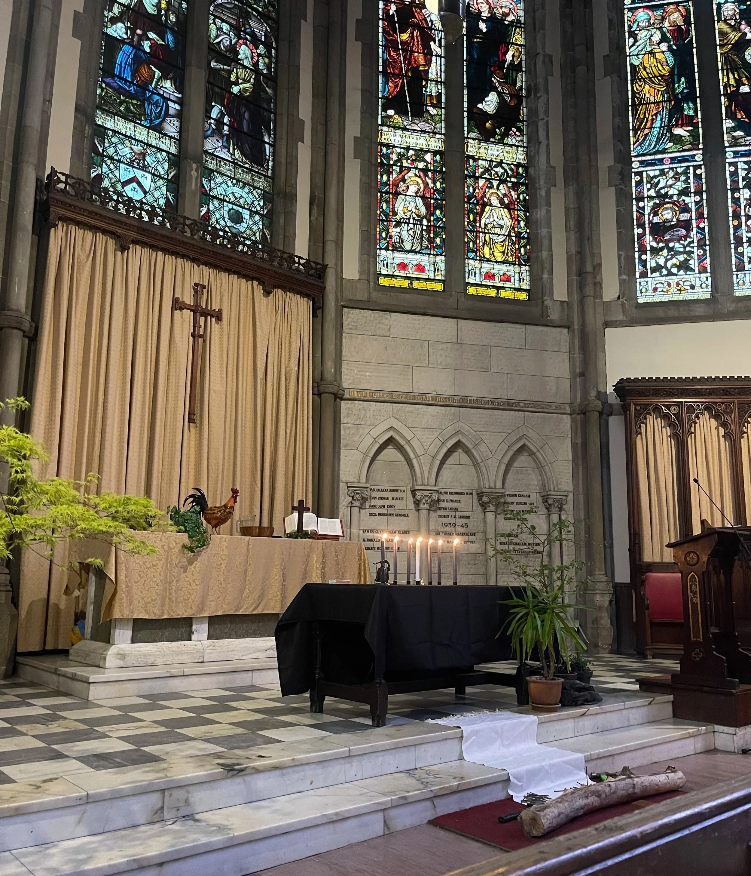 Inside Kelvinside Hillhead, the altar with candles, plants, and a white cloth down the steps, stained glass windows with religious figures, and a large wooden cross. A photo taken from one of the Easter services in 2025.