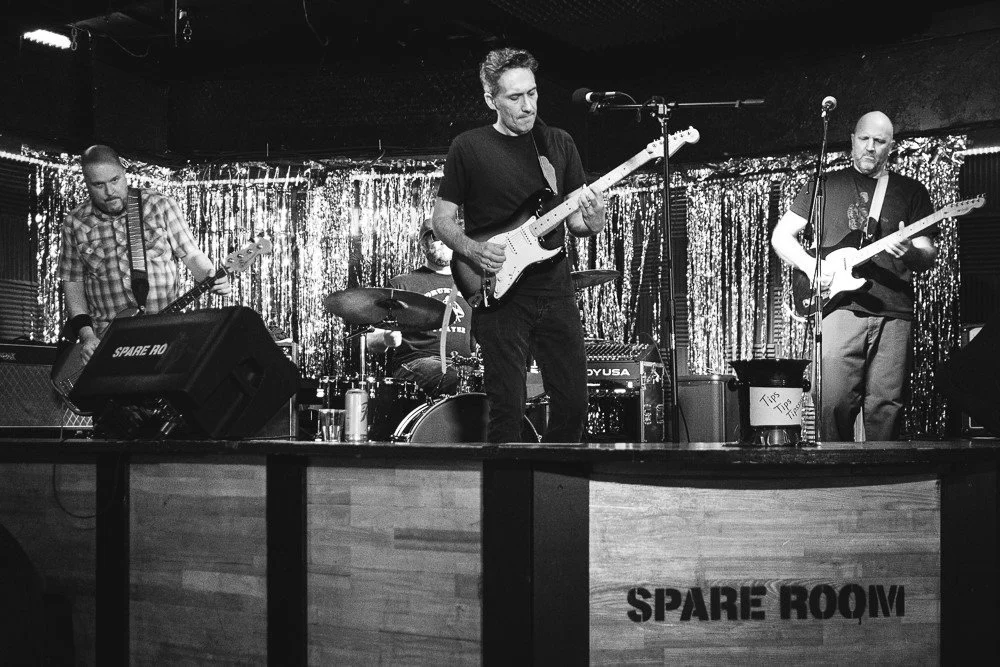 A band of four musicians performing on a stage with a sparkly curtain backdrop. One person plays bass guitar, another plays drums, and two others play electric guitars. There is a sign that says "Spare RQVM" and a tip jar on the stage.