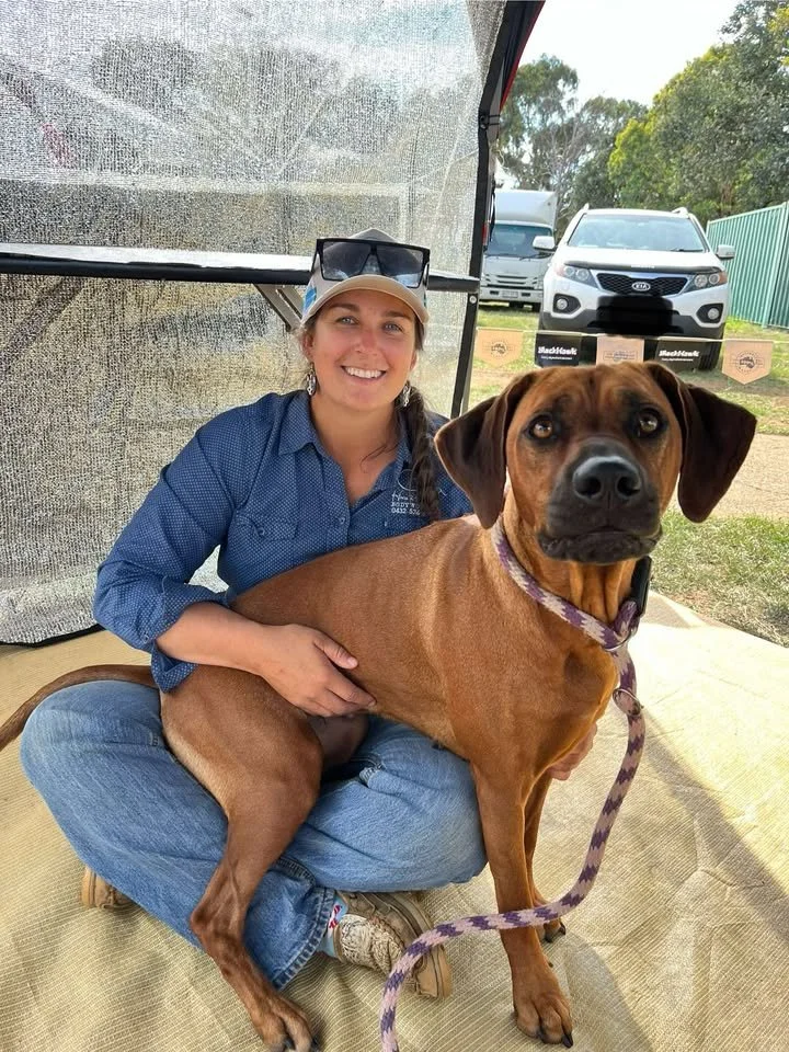 A woman with a smile sitting cross-legged on the ground, holding a large brown dog on her lap. They are outside with cars and trees in the background.