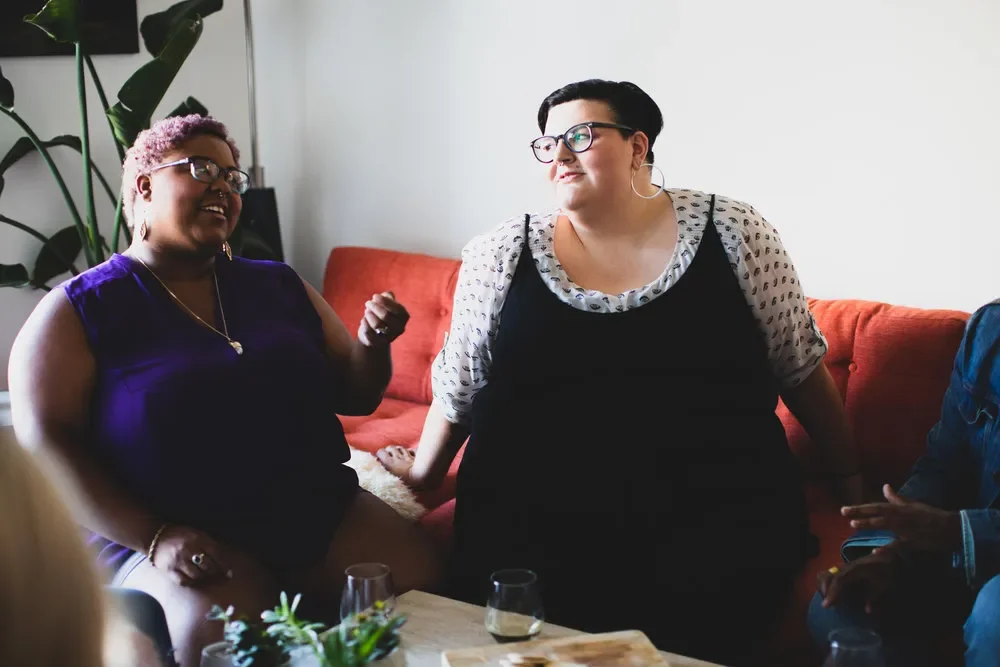 Three women talking and smiling in a living room with a red couch and green plants.