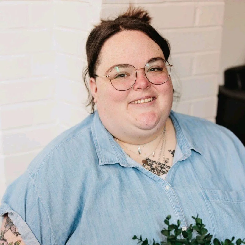 A person with short dark hair, glasses, and a smile, wearing a light blue denim shirt, standing in front of a white brick wall, holding a small plant with green leaves.