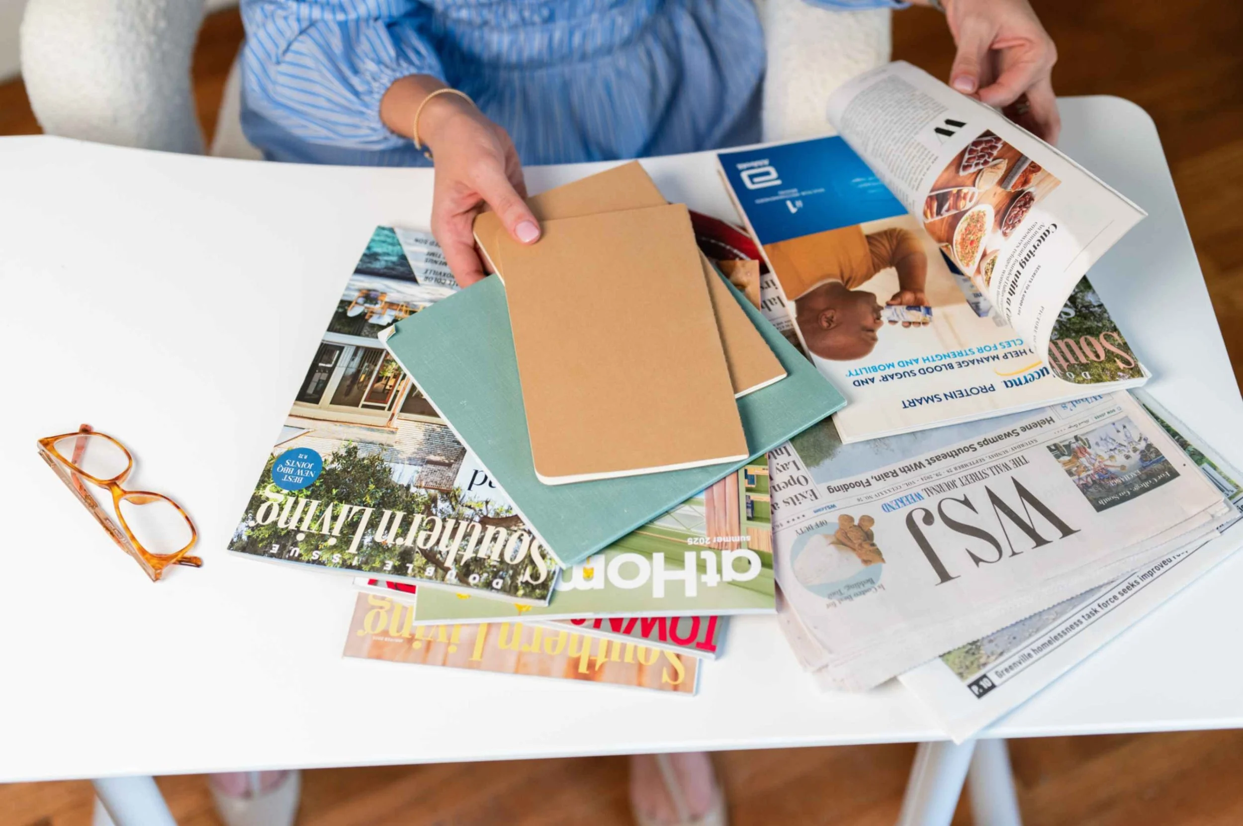 Person sitting at a white table, looking through magazines and pamphlets, with orange glasses on the table.