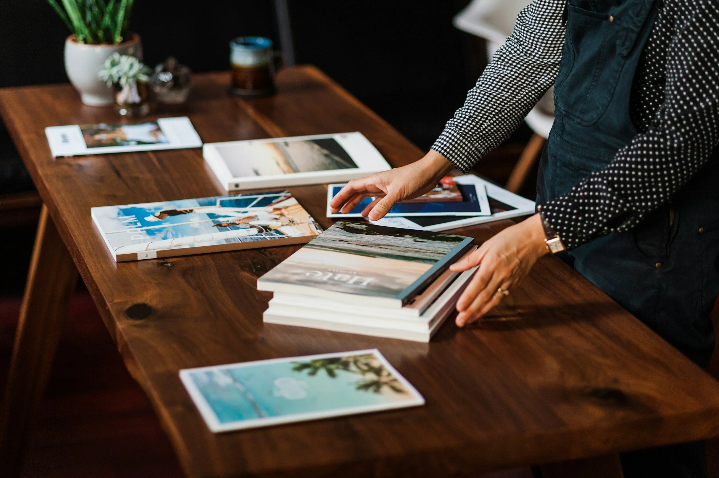 Person arranging travel brochures and magazines on a wooden table, with a plant and cups in the background.