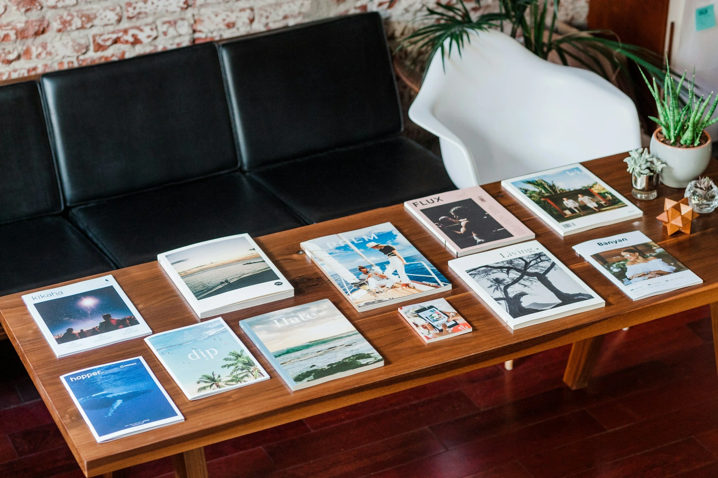 A wooden table displaying various magazines and pamphlets, with a black sofa, white chair, and plants in the background.