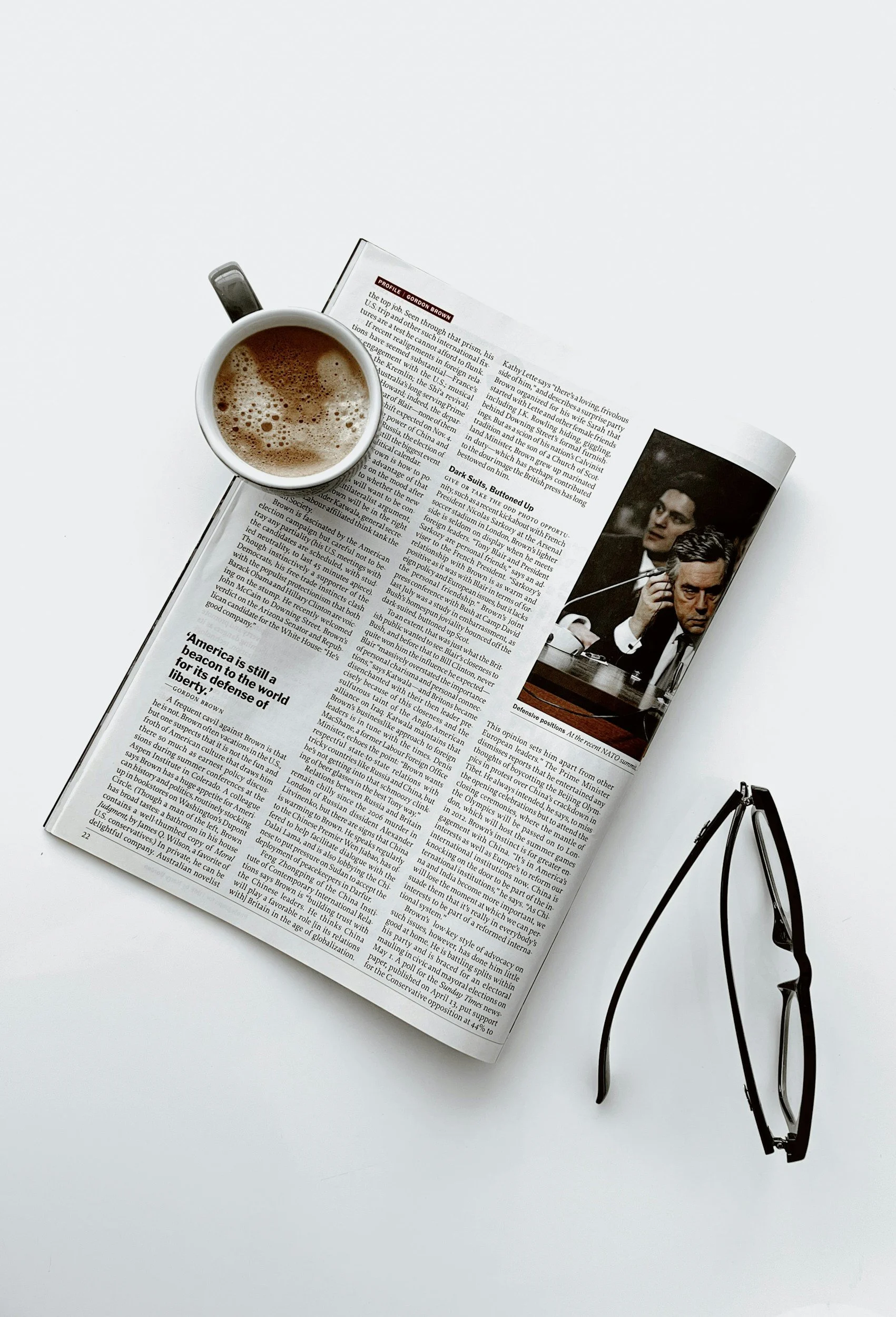 A cup of coffee on an open newspaper with a pair of glasses on a white surface.