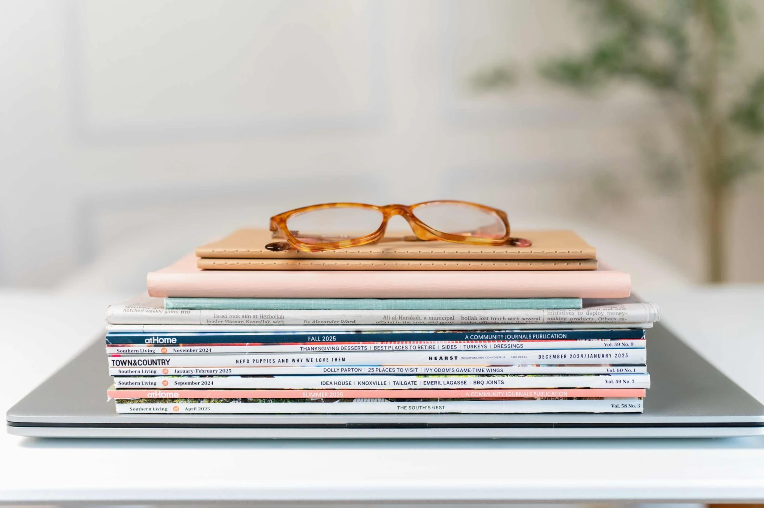 Stack of magazines and notebooks with a pair of orange tortoiseshell glasses on top, sitting on a silver laptop on a white surface.