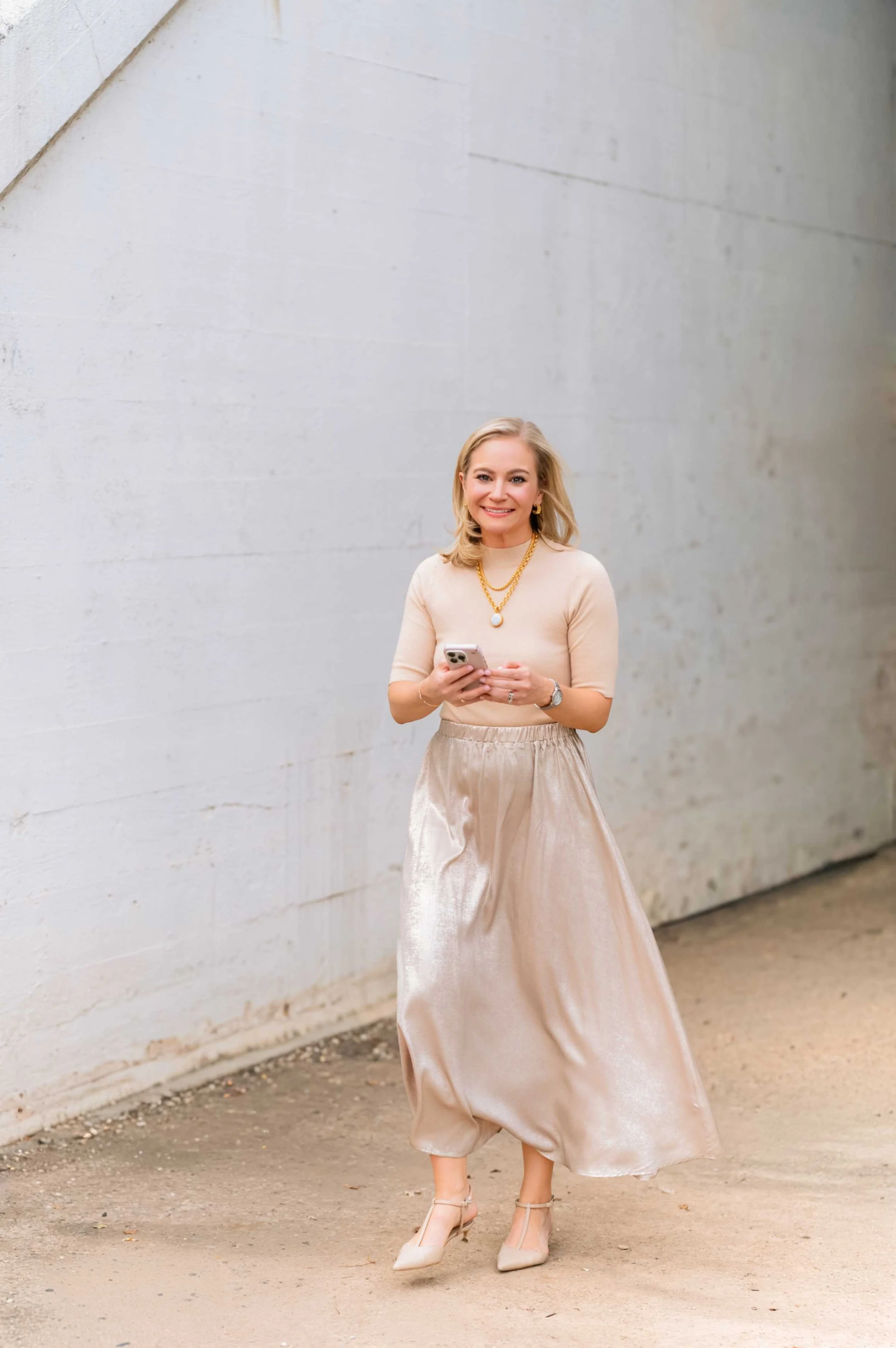 A woman smiling while holding a phone, wearing a beige top, a shiny beige skirt, and beige heels, standing against a white wall.