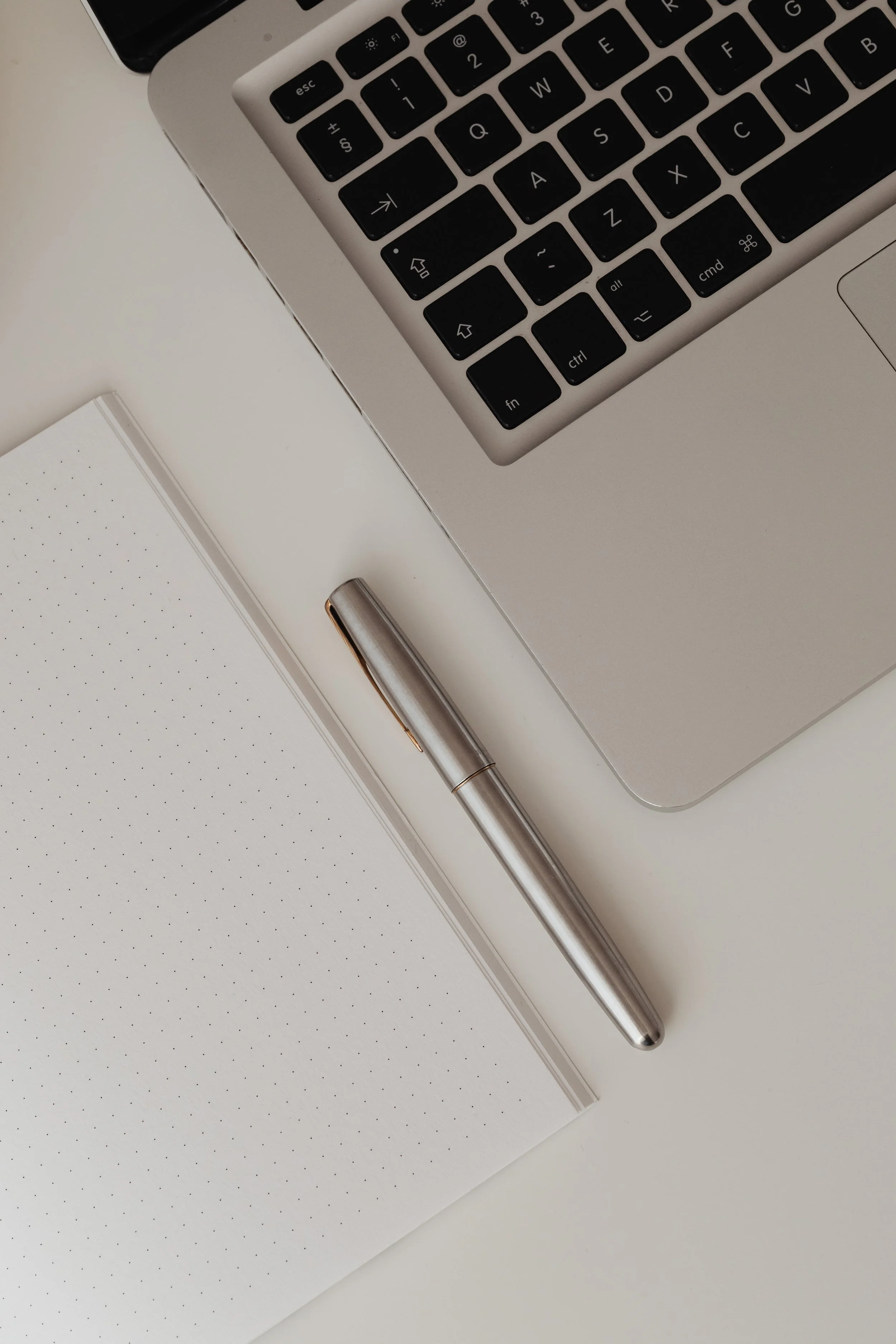 Close-up of a silver laptop keyboard, a silver pen, and a dotted notebook on a white desk.