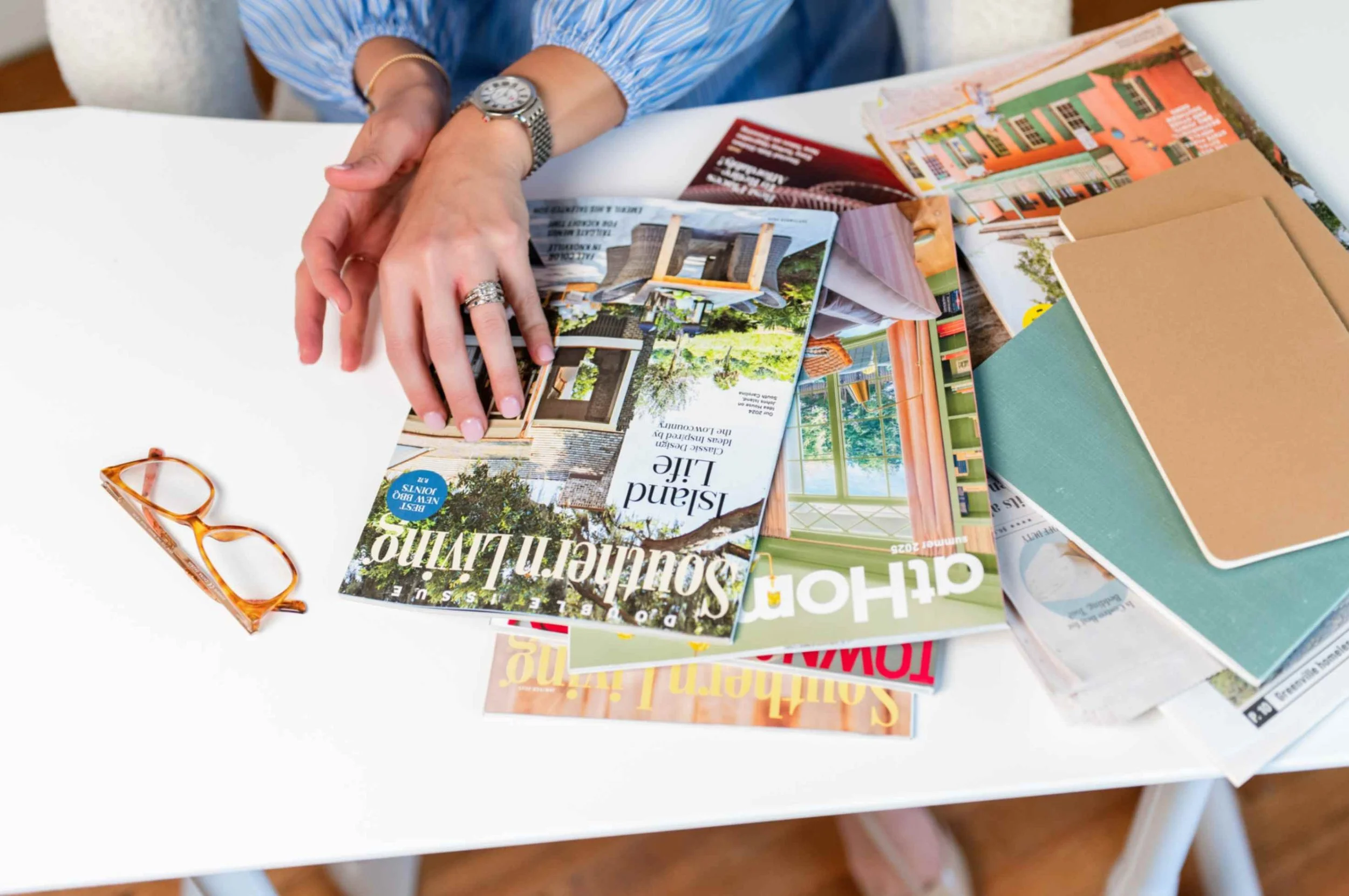 A person's hands with rings and a watch, resting on a table with scattered real estate magazines, brochures, and orange glasses.