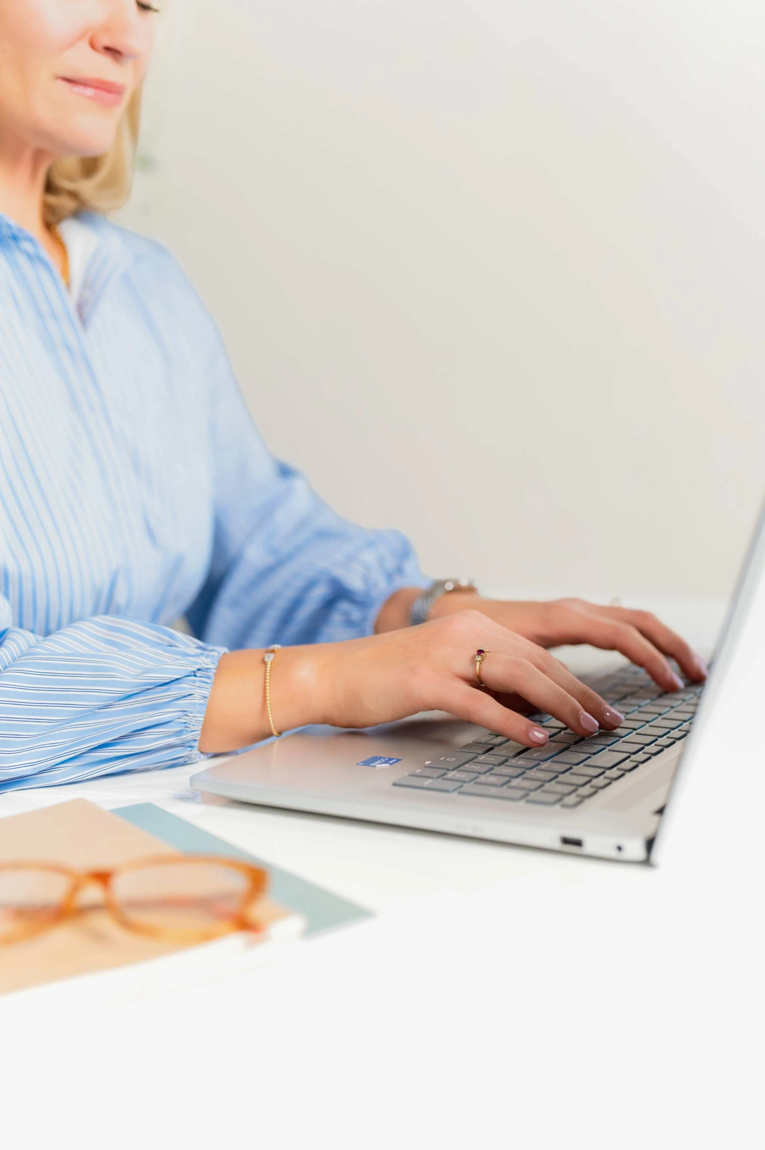 Woman typing on a silver laptop with glasses and papers on desk.