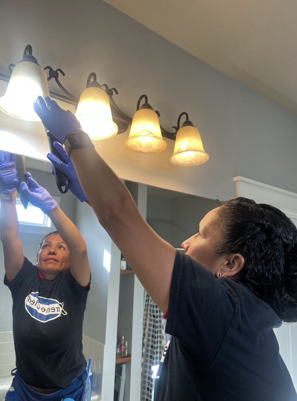 Two women are working together to replace a light fixture in a bathroom. One is holding a tool while the other is assisting, both wearing gloves.
