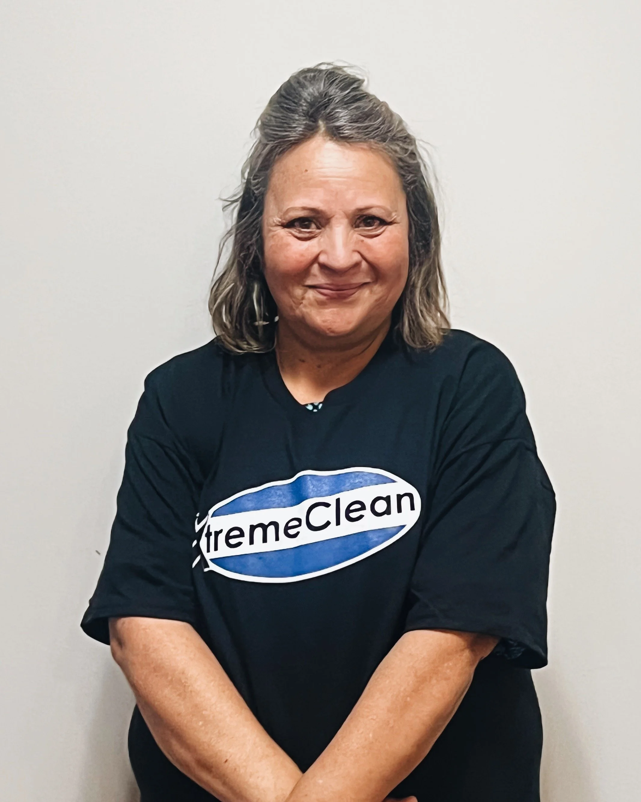 A woman with shoulder-length gray hair smiling, wearing a black 'ExtremeClean' T-shirt, standing against a plain white wall.