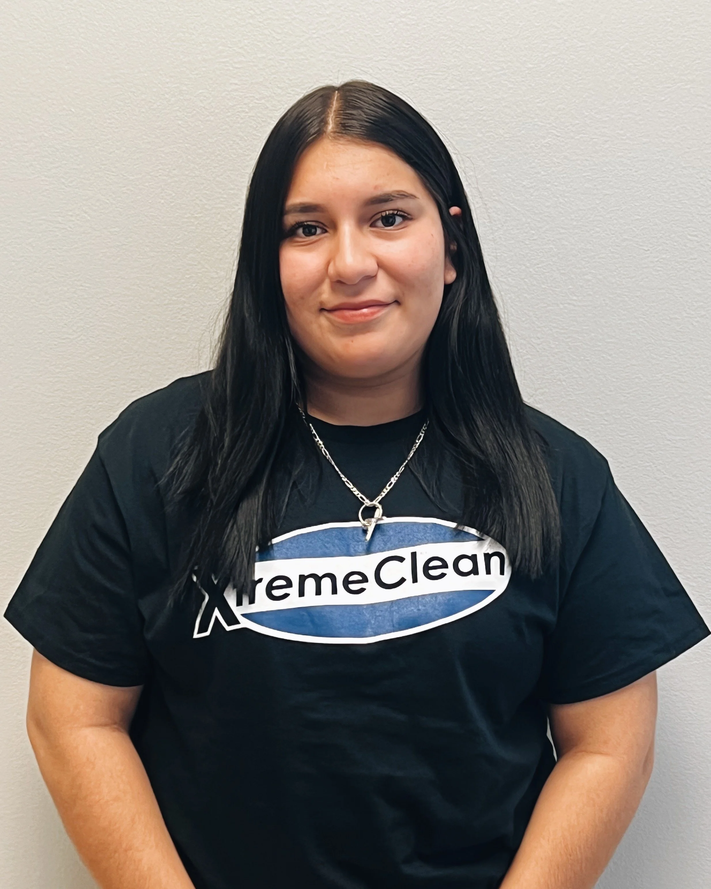 Young woman with long black hair wearing a black T-shirt with the words 'XtremeClean' and a necklace, standing against a plain light-colored wall.