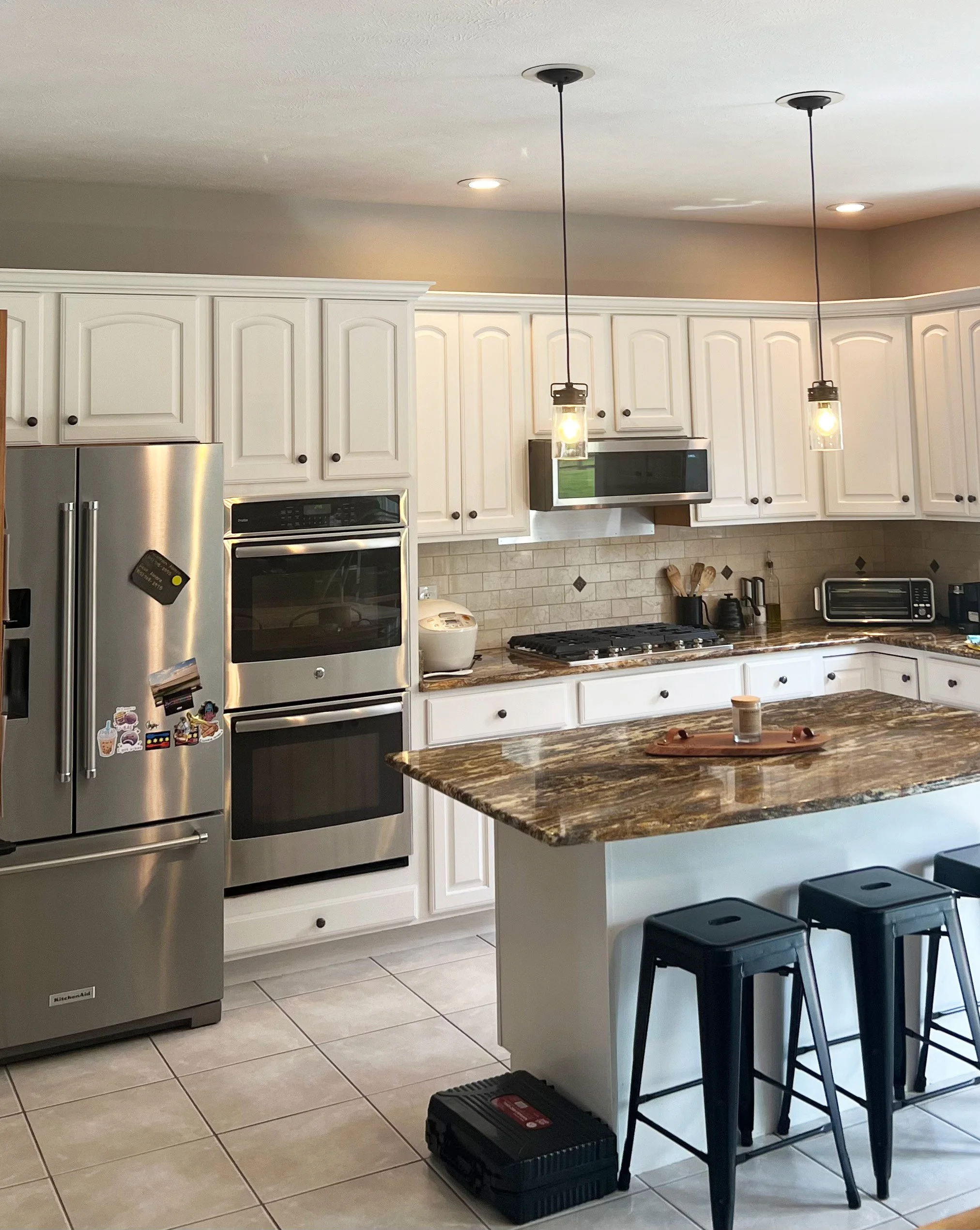 Modern kitchen with white cabinets, stainless steel appliances, an island with a marble top, black bar stools, pendant lights, and tiled flooring.