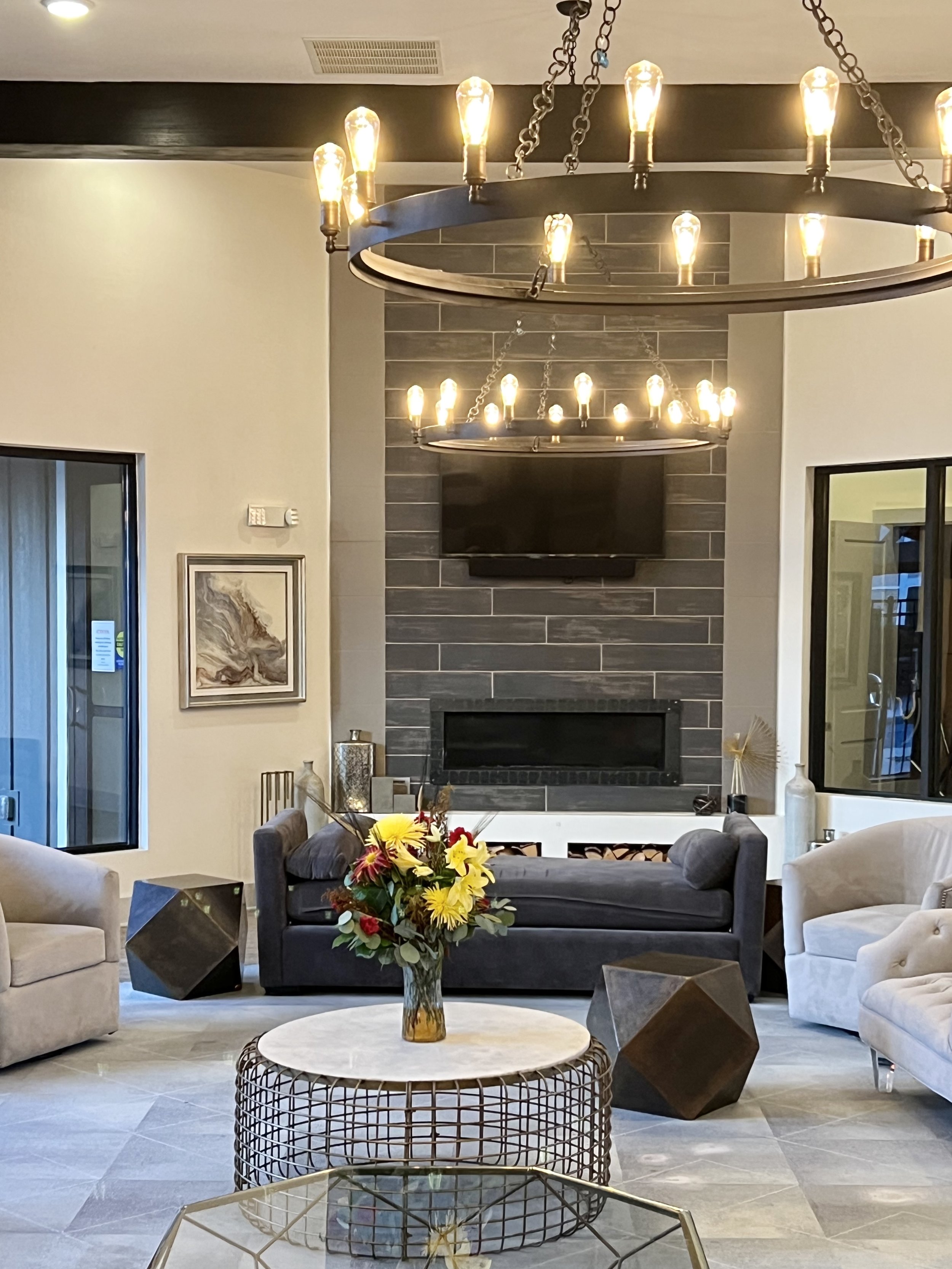 A modern living room with a fireplace, mounted television, and a chandelier. There are beige, gray, and dark gray sofas, a floral arrangement on a round table, and decorative objects.