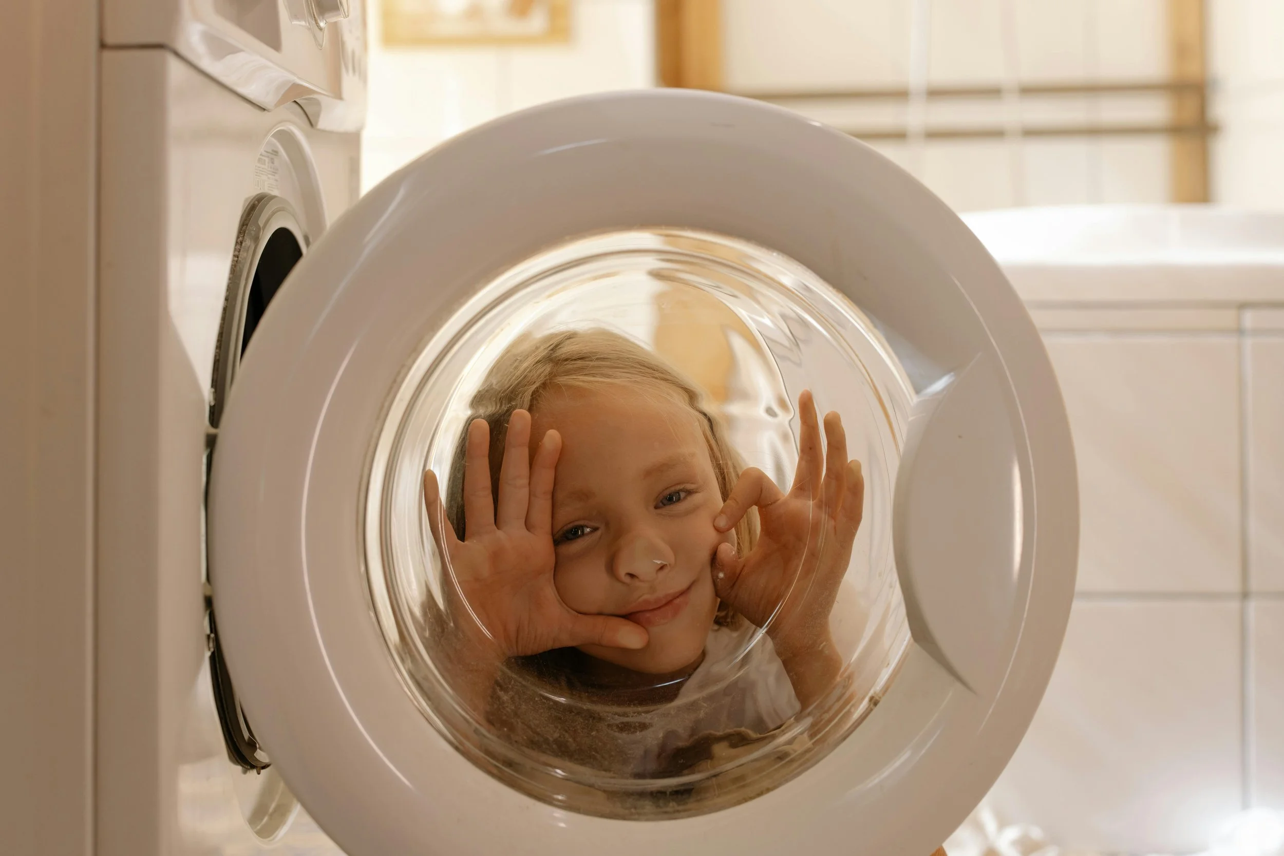 Child peering into a front-loading washing machine with hands on the glass door, smiling.