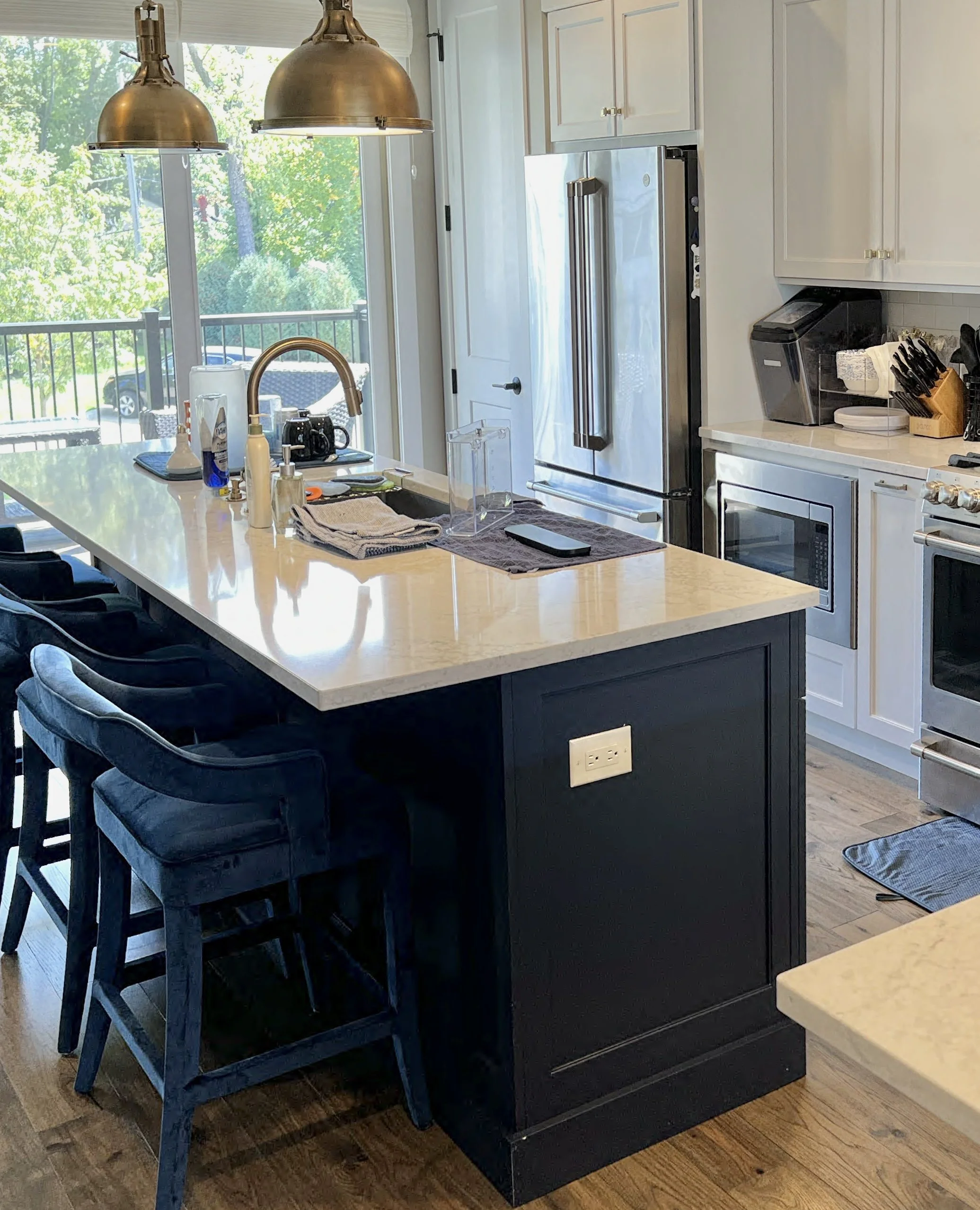 A modern kitchen with a large island, black cabinets, white upper cabinets, stainless steel appliances, and a sliding glass door overlooking a backyard with green trees.