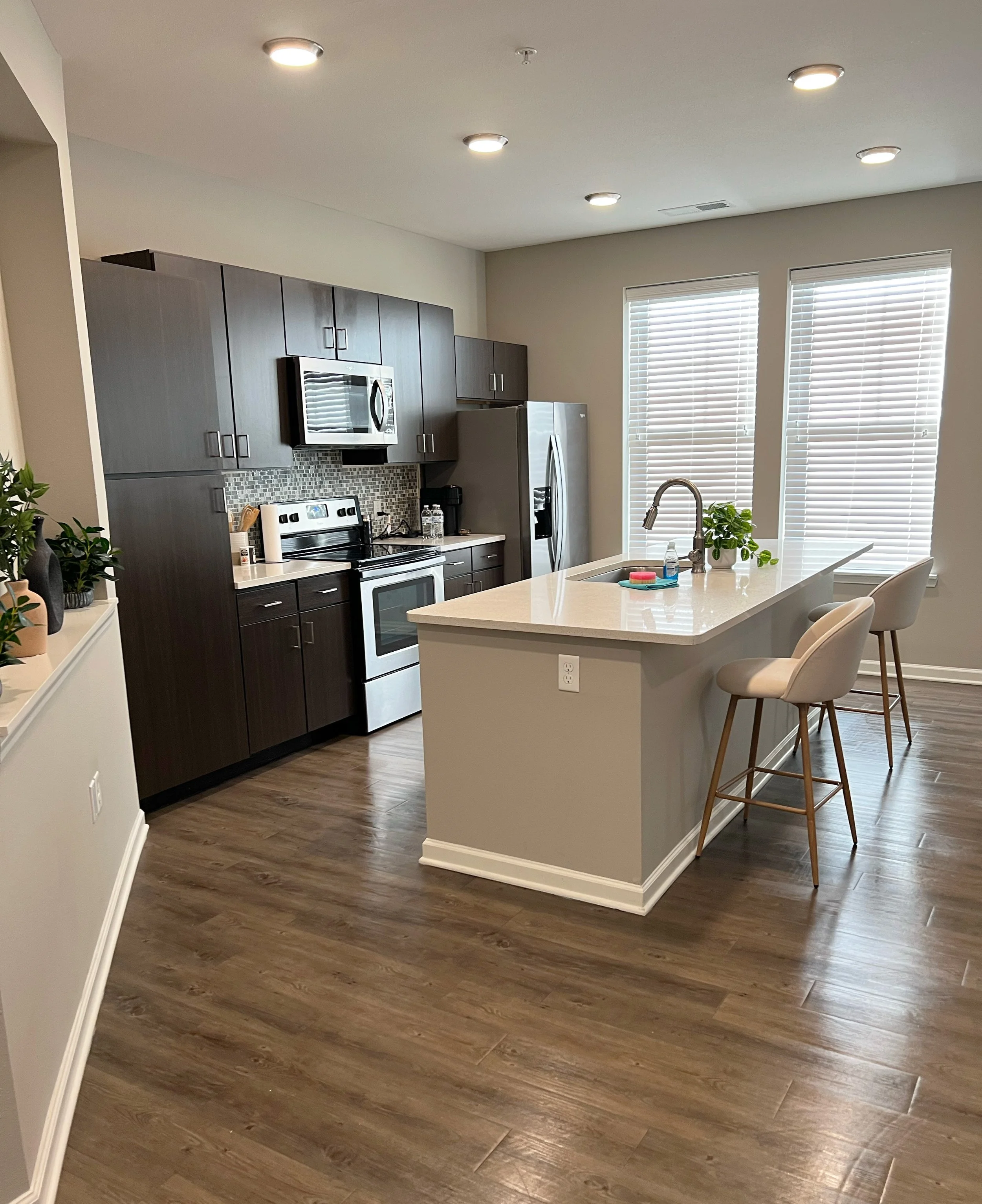 Modern kitchen with dark wooden cabinets, stainless steel appliances, a white kitchen island with beige barstools, and two large windows with white blinds
