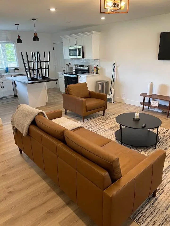 Living room with brown leather sofa and armchair, black round coffee table, TV on wall, and kitchen in background with island, white cabinets, and oven.
