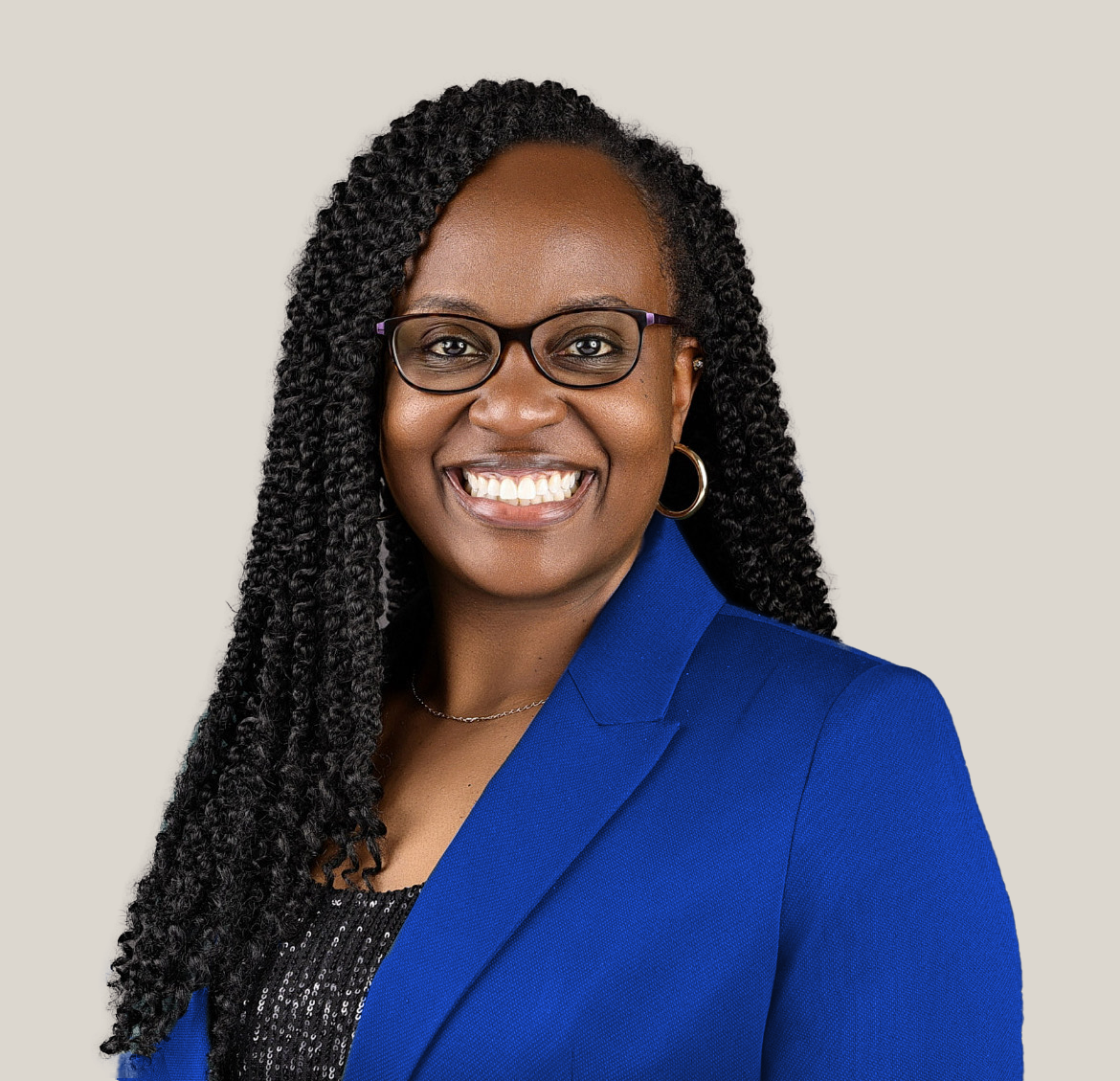 Portrait of a smiling woman with glasses, hoop earrings, wearing a blue blazer and a sequined top against a neutral background.