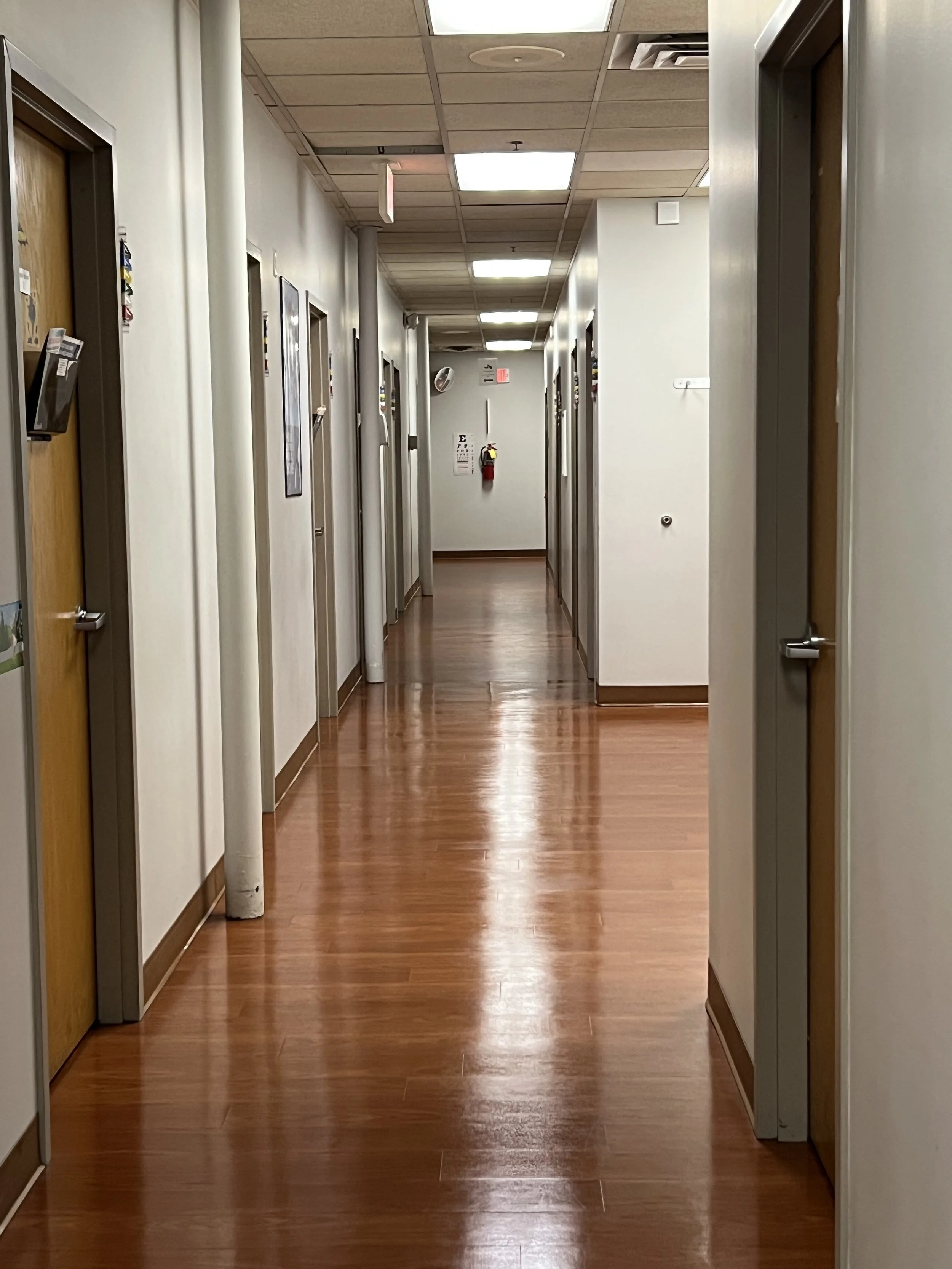 Empty hospital corridor with closed doors on both sides, wood flooring, white walls, ceiling lights, fire extinguisher at the end of the hall.