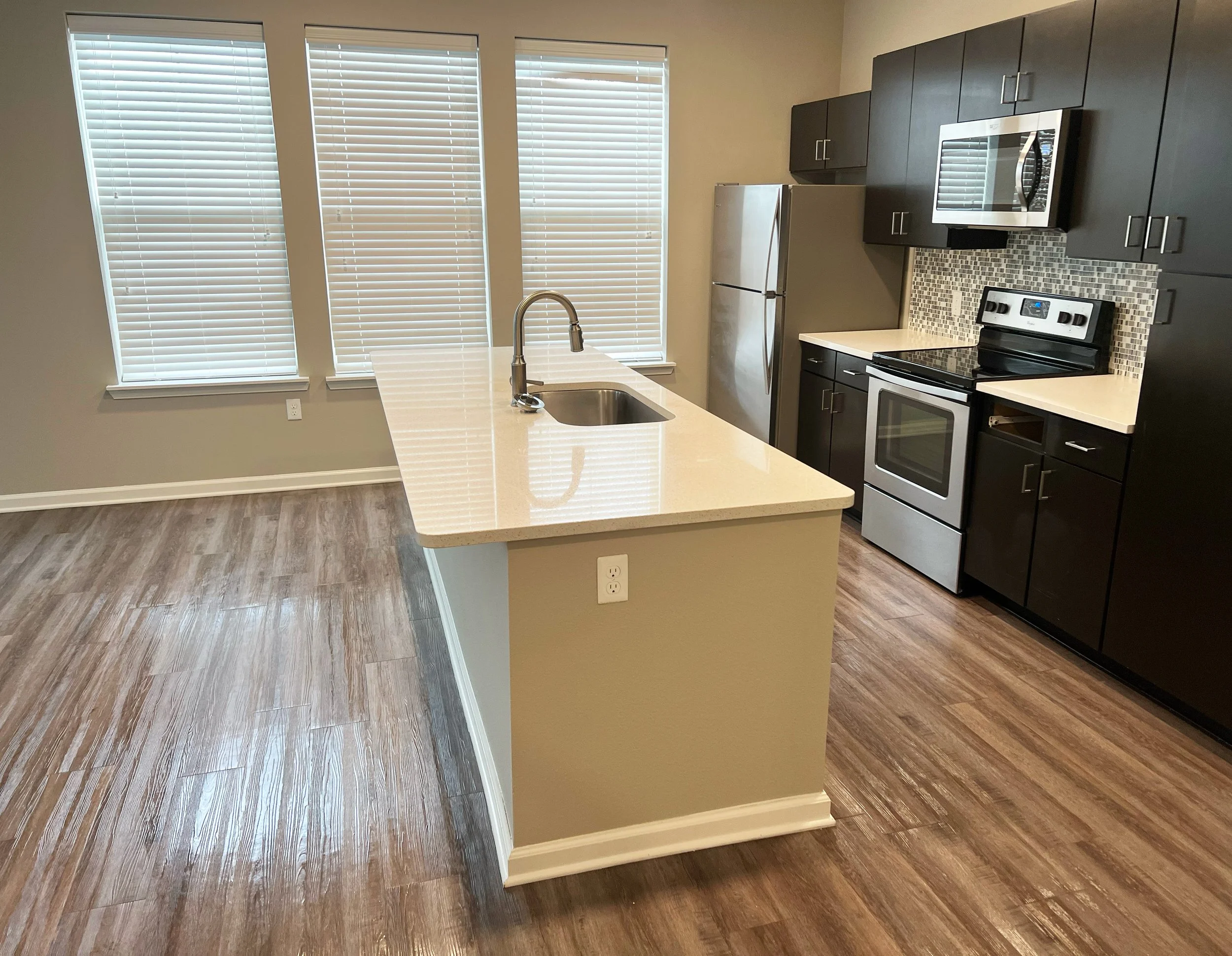 Kitchen with three large windows with blinds, beige walls, an island with a sink, dark cabinets, stainless steel refrigerator, stove, microwave, and wood-look flooring.