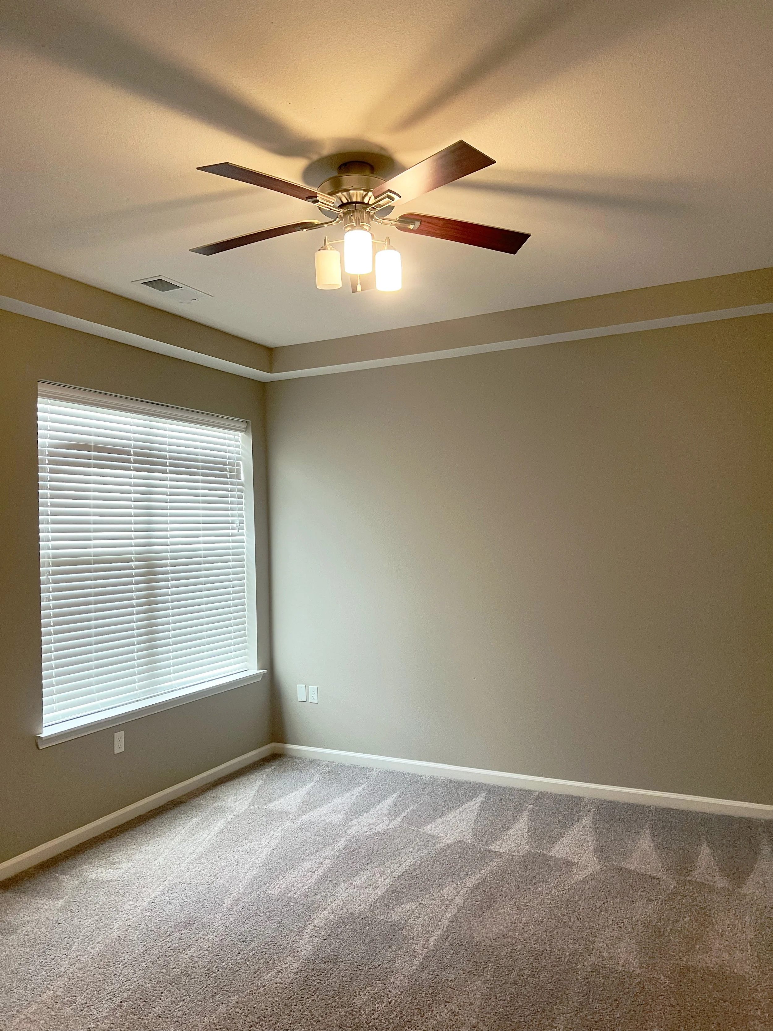 Empty bedroom with a window, beige walls, carpeted floor, and ceiling fan with lights.