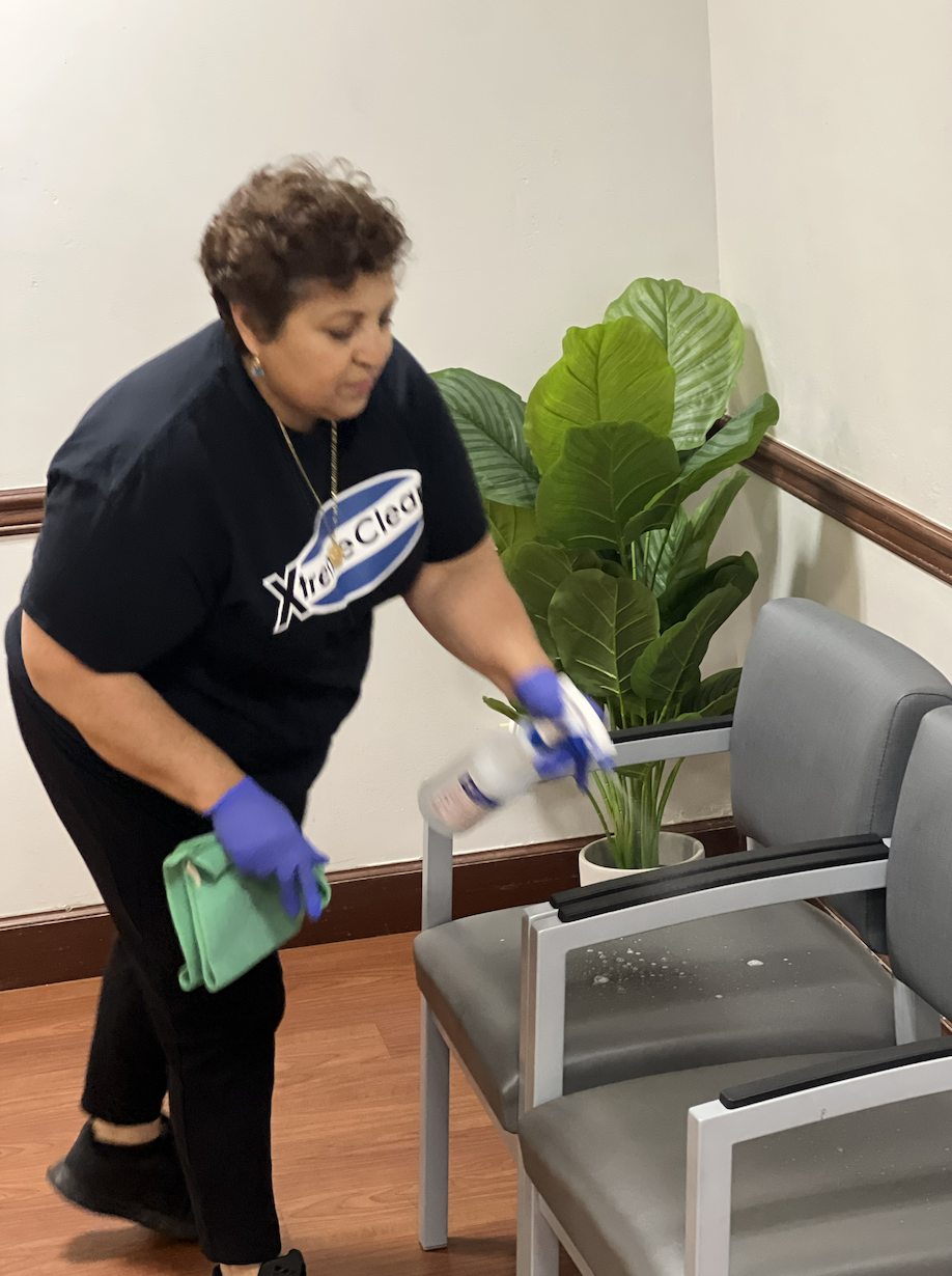 Woman cleaning chairs with spray bottle and cloth in a waiting area, next to a large green plant.